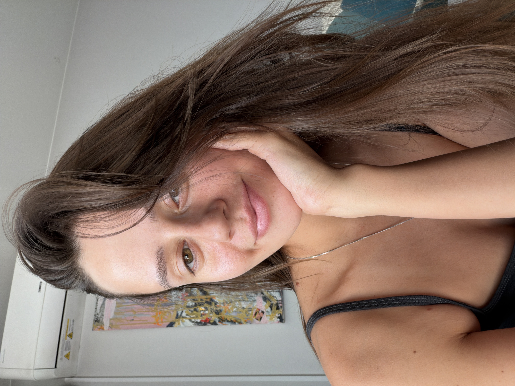 A young woman with long brown hair and brown eyes is looking at the camera with her chin resting on her hand. She is wearing a dark gray tank top and a silver necklace. In the background, there is a white air conditioning unit mounted on a wall, and a colorful abstract painting. The lighting suggests it is daytime. The location is not discernible from the image.