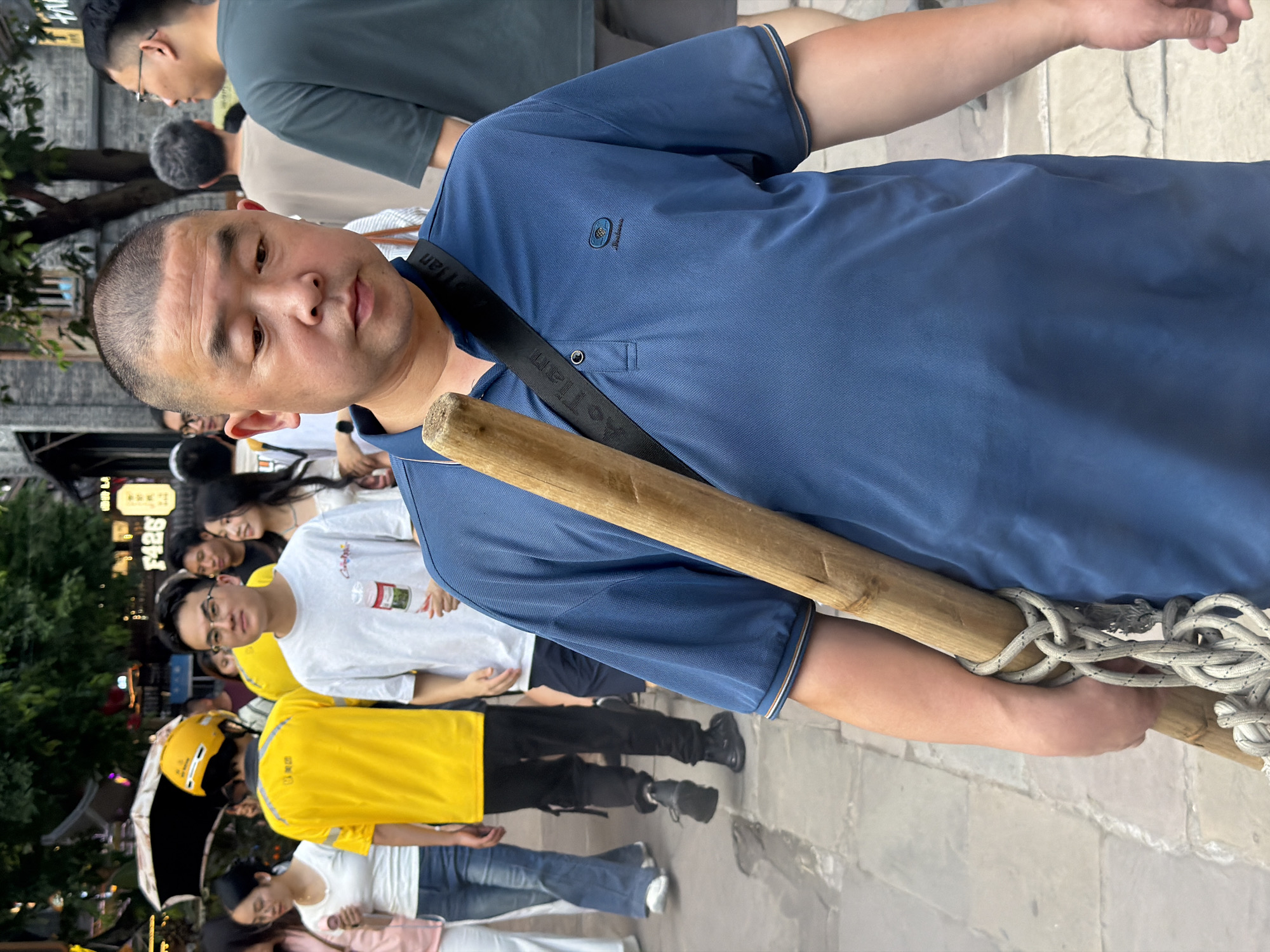 In Yuzhong District, China, a middle-aged East Asian man wearing a blue polo shirt walks on a paved street lined with shops and trees. He carries a thick wooden pole horizontally across his body, secured by a rope that loops around his shoulder and the pole. The man has a shaved head and is looking downwards. Behind him, several other people are walking in the same direction. Among them is a person wearing a bright yellow jacket and a yellow helmet, likely a delivery worker. The environment suggests a bustling urban street with commercial establishments.