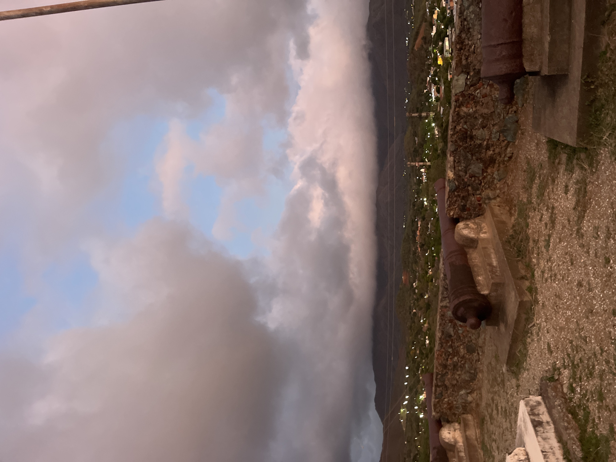 This is a vertical, medium-shot photograph taken at dusk, looking out over a city and mountainous landscape. The foreground features a stone wall, a rocky ground surface, and three large, weathered cannons positioned on concrete mounts. The cannons are dark brown and appear to be made of iron, showing signs of age and rust. Behind the cannons, the stone wall is rough and uneven.
In the middle ground, a town is visible nestled in a valley. Lights from houses and buildings twinkle across the landscape, suggesting it is twilight. The town is surrounded by rolling hills and the distant, dark silhouettes of mountains, which are partially obscured by a dense layer of clouds.
The sky above is a dramatic mix of dark gray and pinkish-purple clouds, with patches of pale blue sky peeking through. A tall, thin, wooden pole is visible on the right edge of the frame, extending upwards into the cloudy sky.
The scene evokes a historical and somewhat somber atmosphere, likely captured from a vantage point such as Fortín de San Carlos Borromeo, a historical fort that offers panoramic views of the surrounding area of Santa Ana, Venezuela, including the city and the mountains.