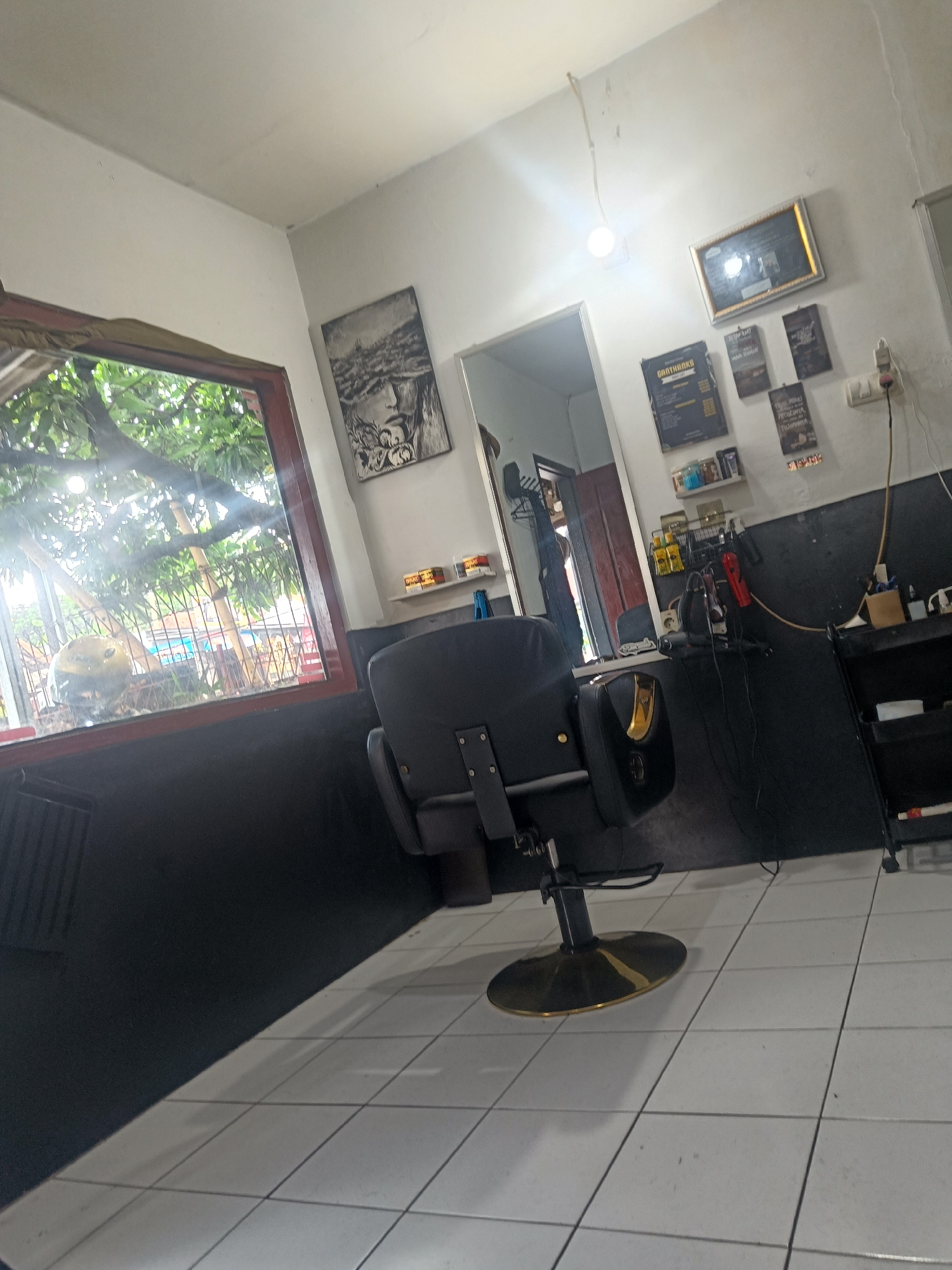 The image depicts a barber shop setting, likely in Cijoho, Indonesia. The foreground features a black barber chair with gold accents positioned on a tiled floor. A window on the left offers a view of greenery and a glimpse of the neighborhood. The walls are painted white and black, adorned with framed pictures, posters, and shelves displaying barber supplies. A mirror reflects the interior of the shop. Tools and equipment are arranged on a shelf behind the chair, suggesting an active workspace. The overall impression is of a functional and well-lit barber shop.