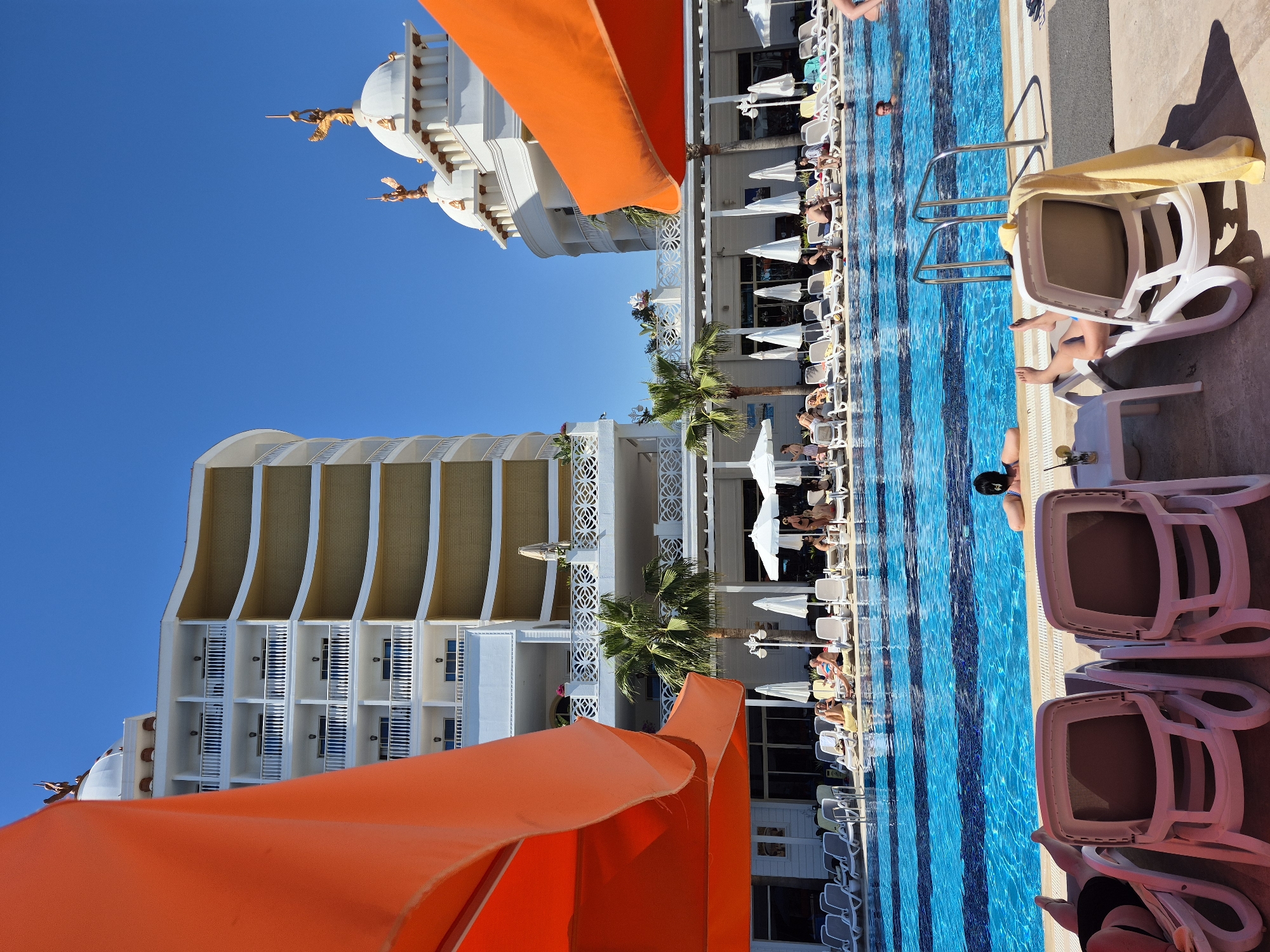 The image captures a scene at a resort in Alanya, Turkey. The view features a blue-tiled swimming pool, lined with white lounge chairs and white umbrellas. People are seen relaxing in the chairs and swimming in the pool. In the foreground, there are bright orange sunshades. The backdrop includes tall, white resort buildings with decorative details and golden statues on the roof. The sky is clear and blue.