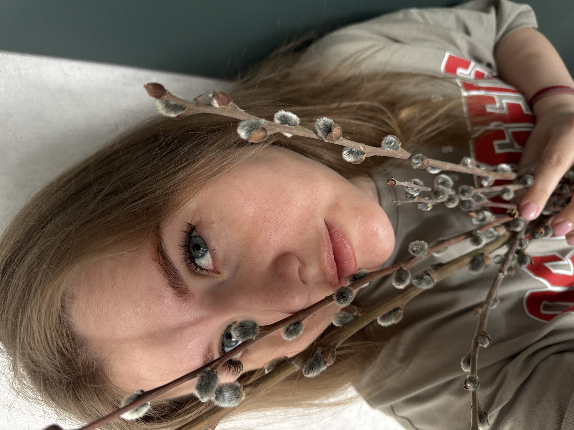 A fair-skinned young woman with light blonde hair and blue eyes lies on a white surface with a dark green background behind her. She holds several branches of pussy willow in her right hand, positioning them near her face. She is wearing a grey t-shirt with red lettering. The focus is on her face and the branches, which obscures most of her clothing. The image is taken from a high angle. The location is not discernible from the image and could not be related to the specified city.