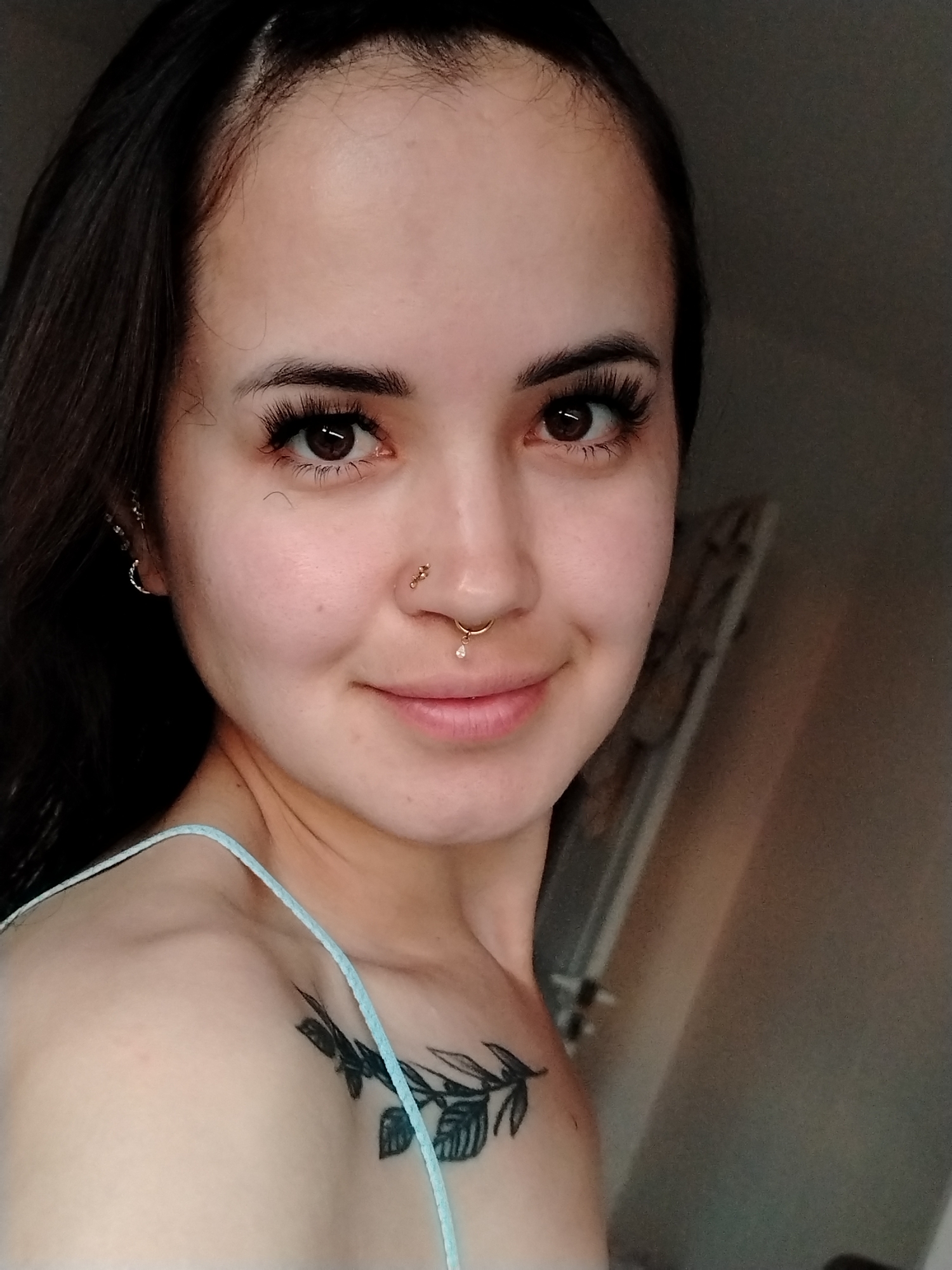 A medium shot captures a young woman with dark hair and brown eyes. She is looking directly at the camera with a slight smile. Her makeup includes noticeable false eyelashes and lip gloss. She has piercings on her nose and septum, and an earring in her left ear. A tattoo of branches and leaves is visible on her left shoulder and chest. She is wearing a light blue strap. The background is a neutral, out-of-focus gray wall. The scene appears to be indoors, with artificial lighting. The location is identified as Noviy, Russia.