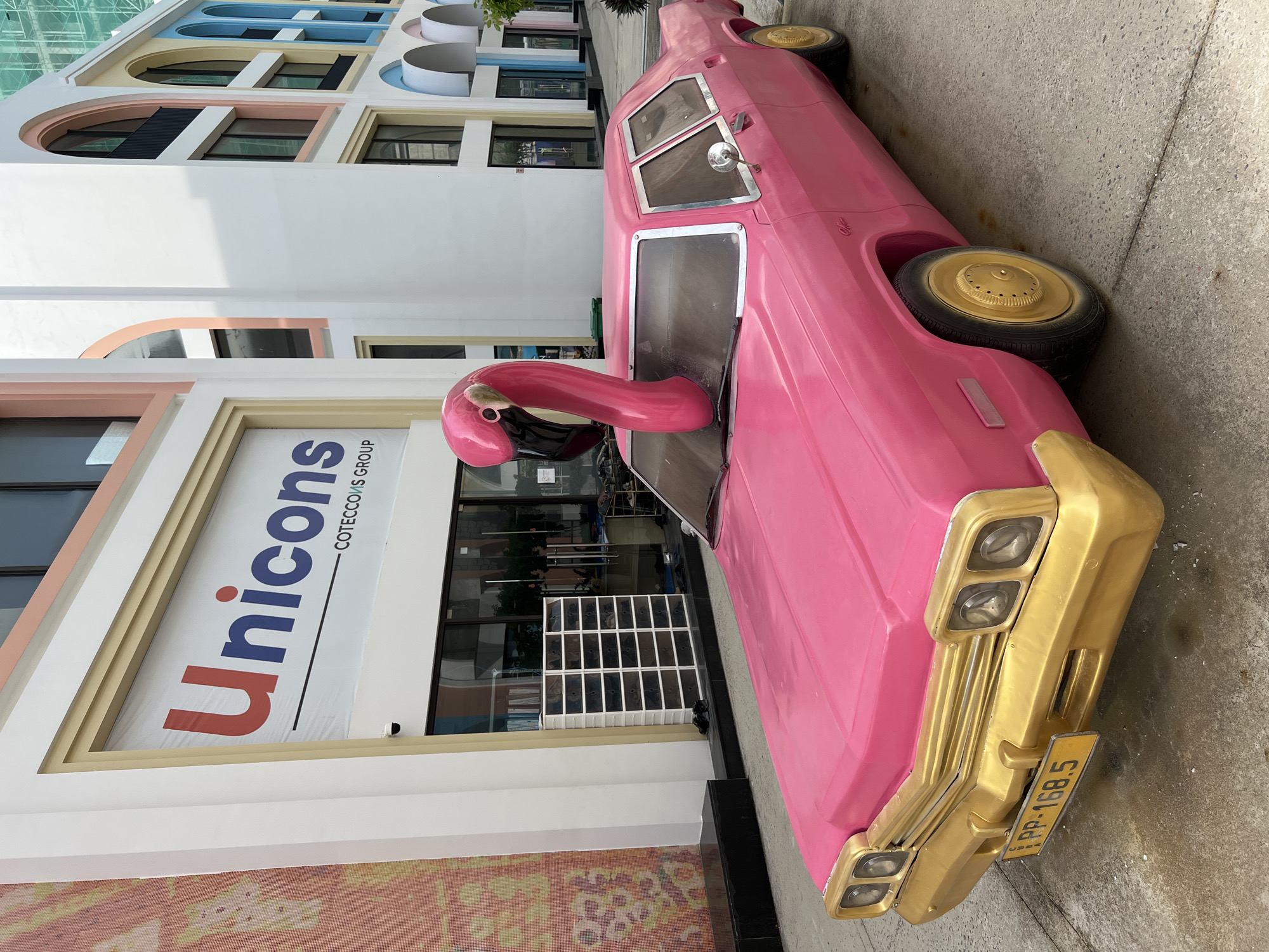 A pink classic car, adorned with gold trim and wheels, is parked on a paved surface in front of a modern building. A large pink flamingo sculpture emerges from the car's hood, its neck bent at an angle. The building features a large sign with blue and red lettering that reads "Unicons" and "COTECCONS GROUP". The building's exterior is decorated with colorful arched windows and a patterned mosaic wall. The scene appears to be a public space, likely a commercial area or an attraction. The location is Nha Trang, Vietnam.