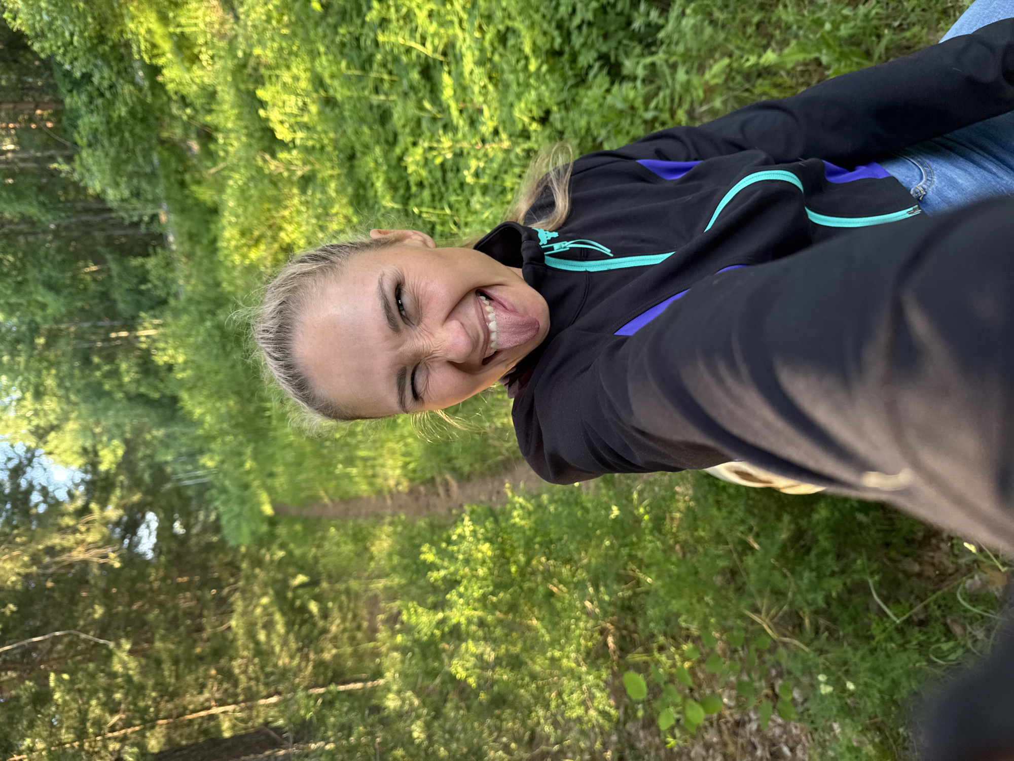 A woman, with blonde hair pulled back in a ponytail, sticks her tongue out at the camera while standing on a path in a forest. She is wearing a black jacket with blue and teal accents and blue jeans. The path is lined with lush green foliage, and trees are visible in the background. The sun is shining, casting a warm light on the scene. The image appears to have been taken in Chaikovsky, Russia, a city known for its natural beauty and forested surroundings.