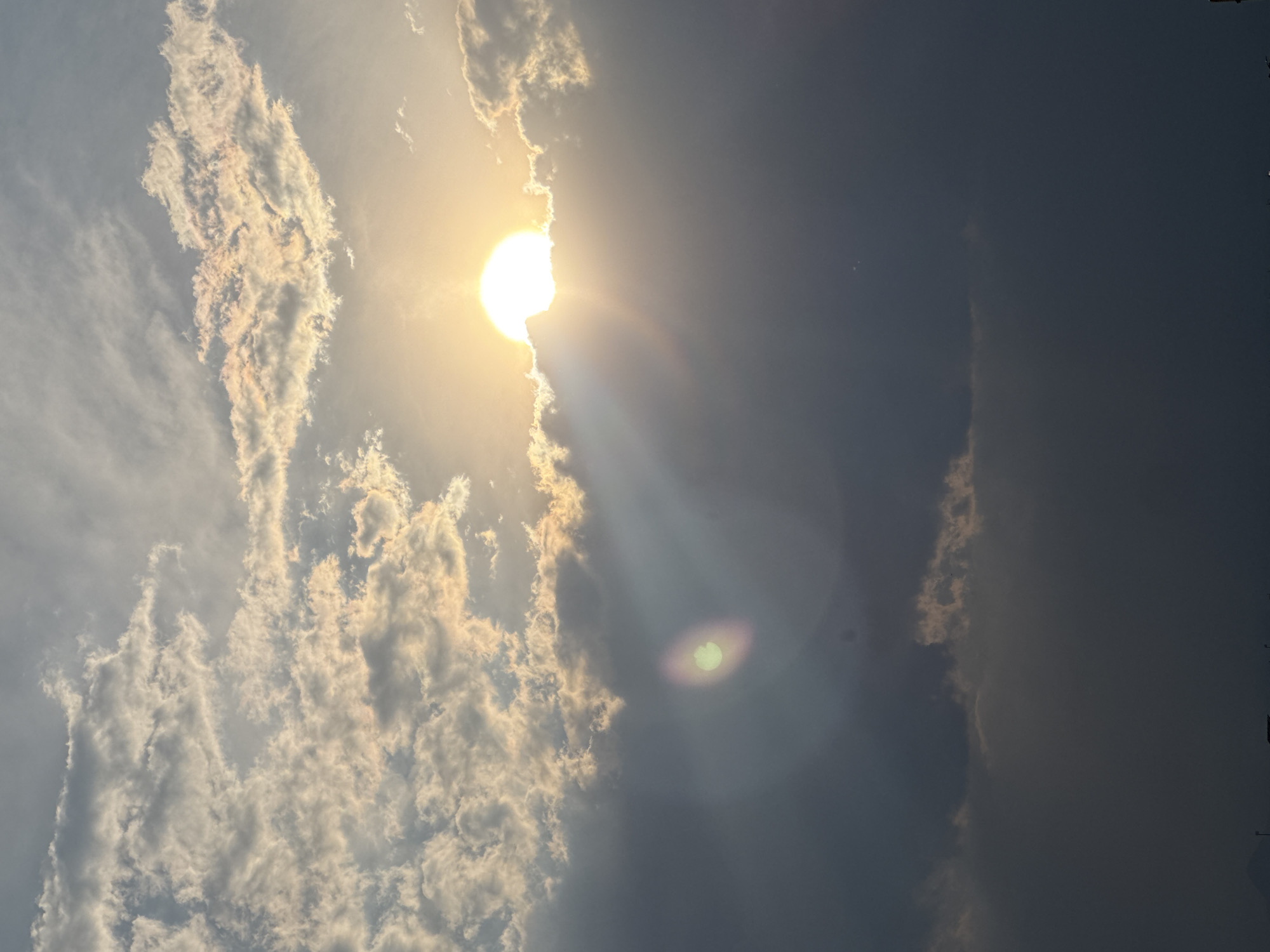The image shows a sky with a bright sun partially obscured by clouds. To the left, there are detailed, puffy clouds reflecting sunlight. To the right, the sky is darker, possibly with heavier cloud cover. A distinct sunbeam is visible. The lens flare is located toward the lower center of the image. The location, San Francisco Ocotlán, Mexico, is not discernible from the visual information provided.