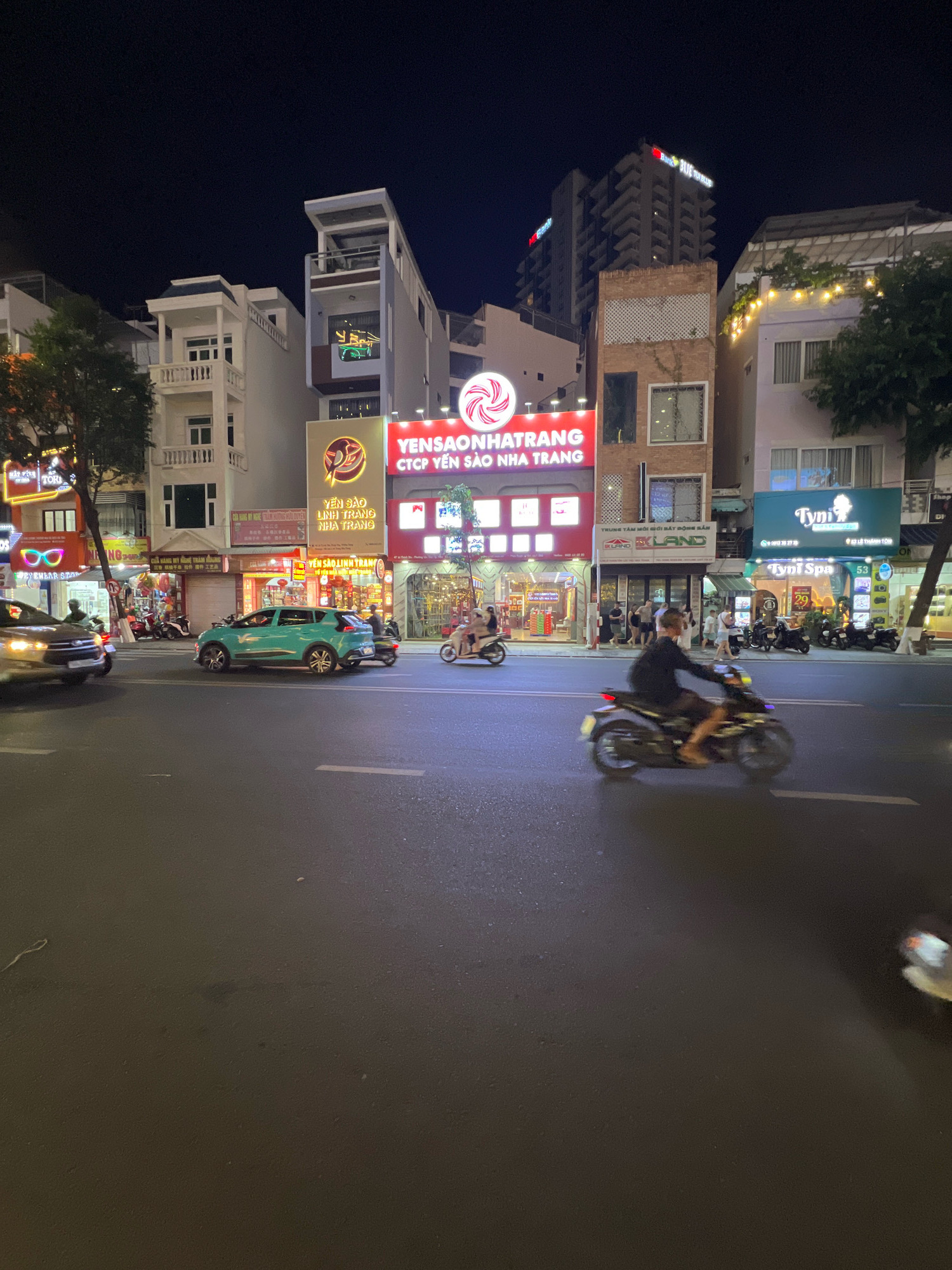 This is a street-level, nighttime photograph taken in Nha Trang, Vietnam. The scene depicts a busy street with several multi-story buildings lining one side. The buildings display various illuminated signs, suggesting commercial establishments. Prominently featured is a large red and white sign with Vietnamese text that reads "YENSAONHATRANG CTCP YẾN SÀO NHA TRANG". Other signs include "TYNI SPA" and "LAND".
The street is paved and shows lane markings. Several vehicles are present, including a turquoise car driving in the street and a blurred motorcycle with a rider in the foreground, indicating movement. Pedestrians are visible on the sidewalk in front of the buildings. Trees and some outdoor lighting, including string lights on one of the buildings, are also part of the environment. The overall atmosphere suggests a vibrant urban setting at night.