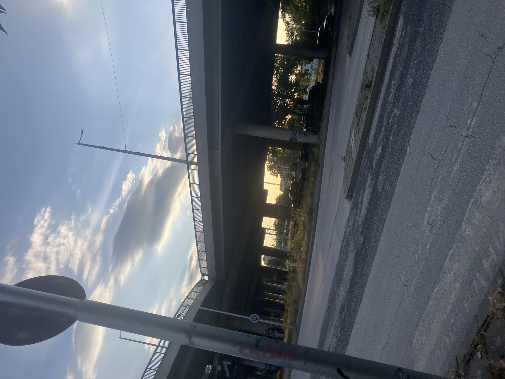An upward-angled shot shows an elevated highway structure in Plovdiv, Bulgaria. The sky is visible with scattered clouds. In the foreground, a road or pathway is visible, marked with alternating light and dark stripes. Vegetation grows along the side of the road. In the middle ground, the highway structure dominates the scene with its supports and concrete construction, obscuring the environment behind. A street lamp stands nearby. The image is taken from a low-angle perspective.