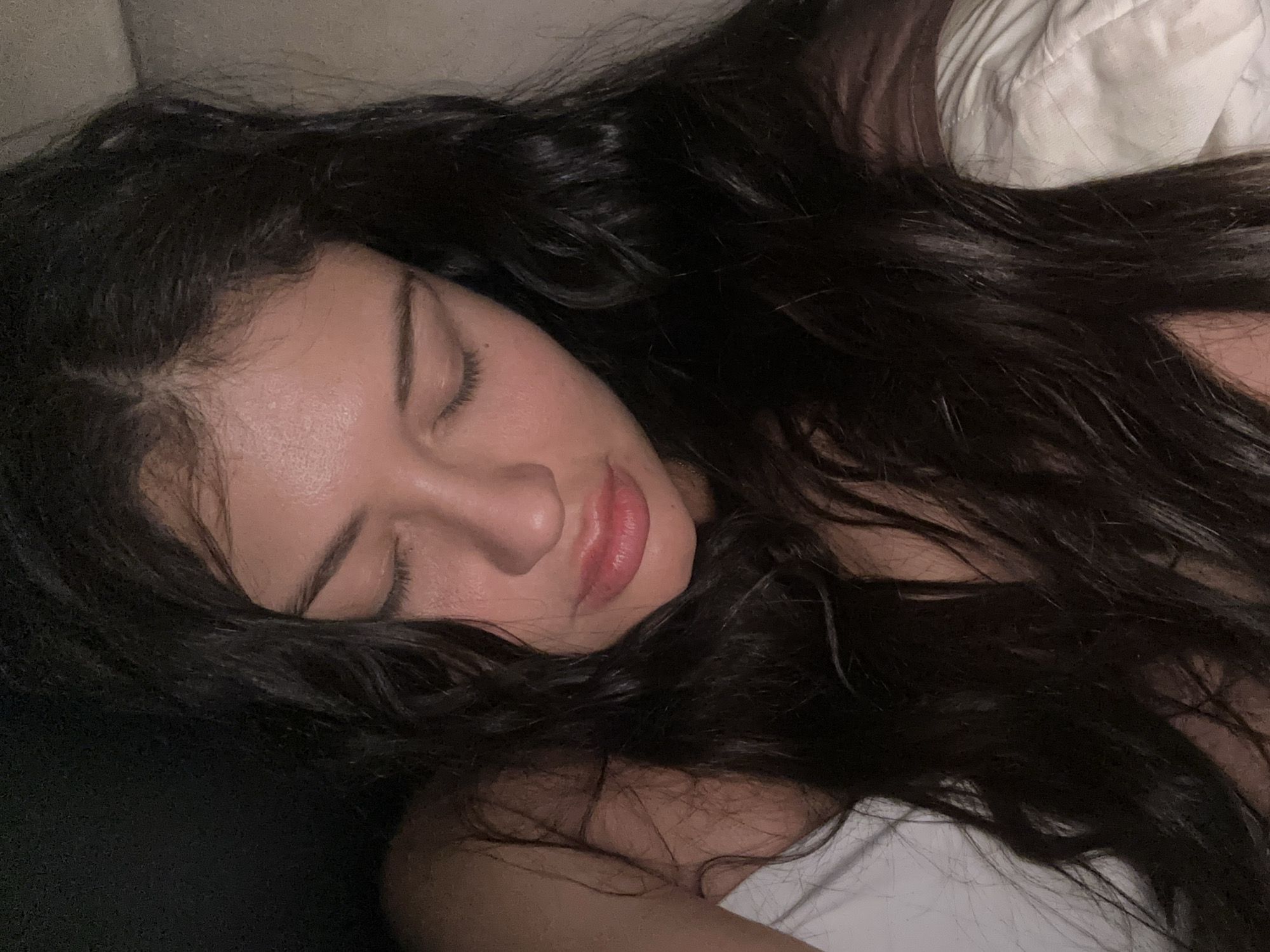 A close-up photograph shows a person with dark, wavy hair lying down with their eyes closed. The person's face is angled towards the camera, and their skin appears smooth. A white pillow is visible in the background, suggesting the person is resting in bed. The lighting is soft and subdued. The image does not provide information related to the city of Khimki, Russia.
