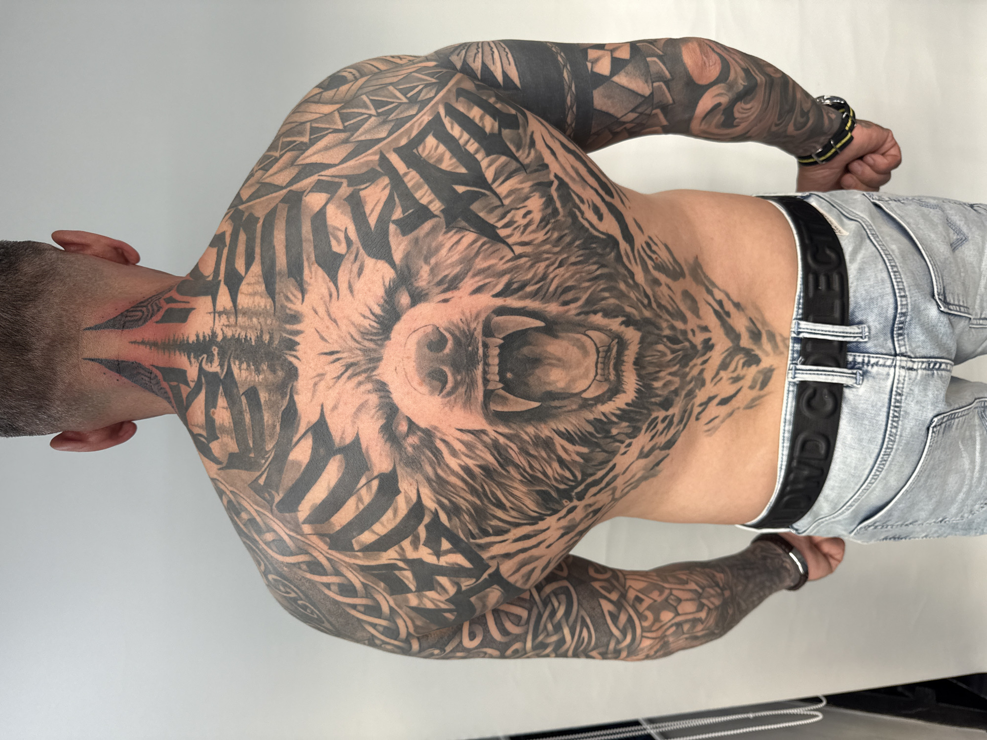 This image captures a male individual from a rear perspective, prominently displaying extensive blackwork tattoos that cover his entire back and both arms. The setting is an indoor space with a plain, light-colored background, suggesting a studio environment in Sofia, Bulgaria.
The central and most striking tattoo on the upper back is a detailed, snarling bear's head with an open mouth and visible teeth, rendered in black and grey realism. This central motif is framed by bold, dark, stylized lettering directly above it, which appears to spell "VALHALLA." Surrounding the bear and the lettering, and extending across the shoulders and upper back, are intricate geometric and tribal patterns, characterized by sharp, angular designs and solid black shapes.
The tattoos continue down both arms, forming full sleeves that seamlessly integrate with the back piece. These arm tattoos feature a diverse mix of Celtic knotwork, swirling motifs, and additional geometric patterns, all executed in a consistent black and grey palette.
The individual has short hair with a clean fade. He is dressed in light blue distressed denim jeans, secured by a black belt. The letters "BWDC" are distinctly visible on the belt. Accessories include a bracelet on his right wrist, featuring black and yellow straps with a metal clasp, and a brown leather bracelet with a silver clasp on his left wrist. The lighting is bright and even, highlighting the intricate details and overall scale of the tattoo artwork. The person is clearly posing to showcase the tattoos, with no other activity taking place.