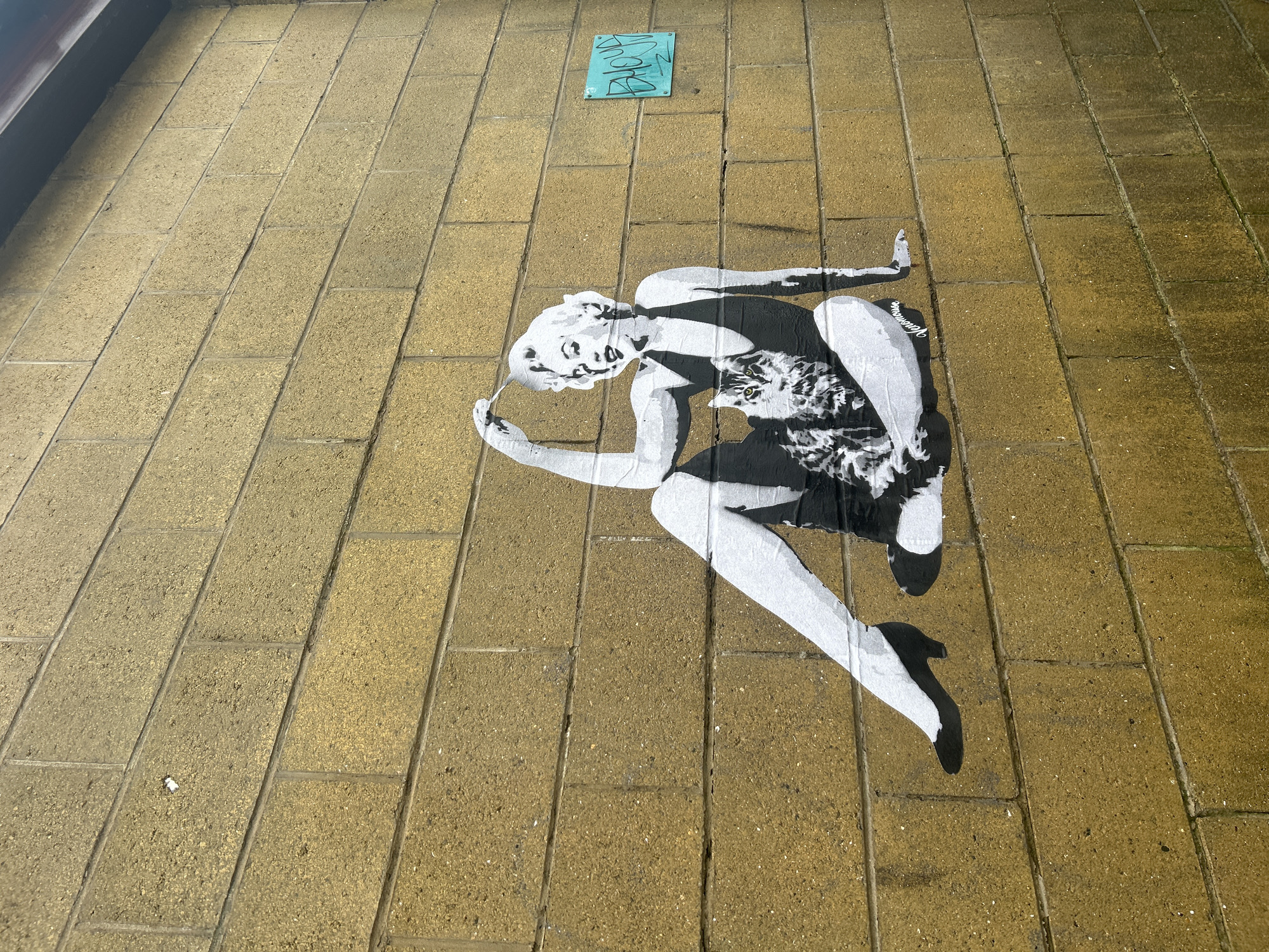 The image is a street scene in Glasgow, United Kingdom. It shows a stenciled artwork on a paved area, likely a sidewalk or pedestrian zone. The artwork is a black and white stencil of a woman in a dress, bending over and seemingly picking something up or interacting with the ground. There is a small, square, blue sign or marker located further up the paved area. The paving stones are rectangular and light brown.
