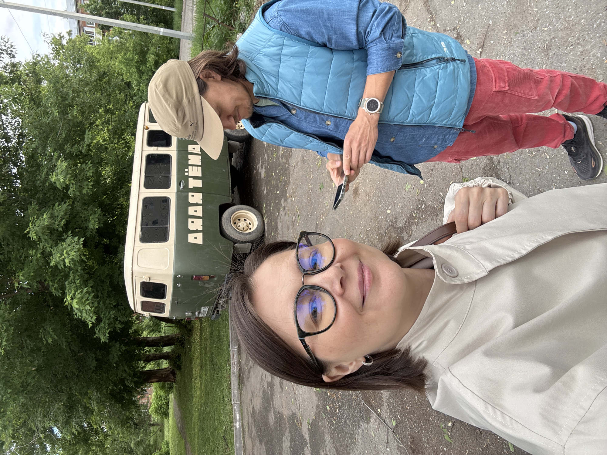 The image shows two individuals and a vintage-style van in what appears to be an urban environment. The person closest to the camera is a woman wearing glasses and a light-colored jacket. She is looking directly at the camera, presumably taking a selfie. Behind her stands a man wearing a light blue vest, a denim shirt, and red pants, and a baseball cap. He is looking at his phone. To their left is a light green and white van with Russian lettering on the side. The van is parked partially on a paved surface and partially on grass. Trees are visible in the background. Based on the writing on the van "Артёма", this could be Izhevsk, Russia.