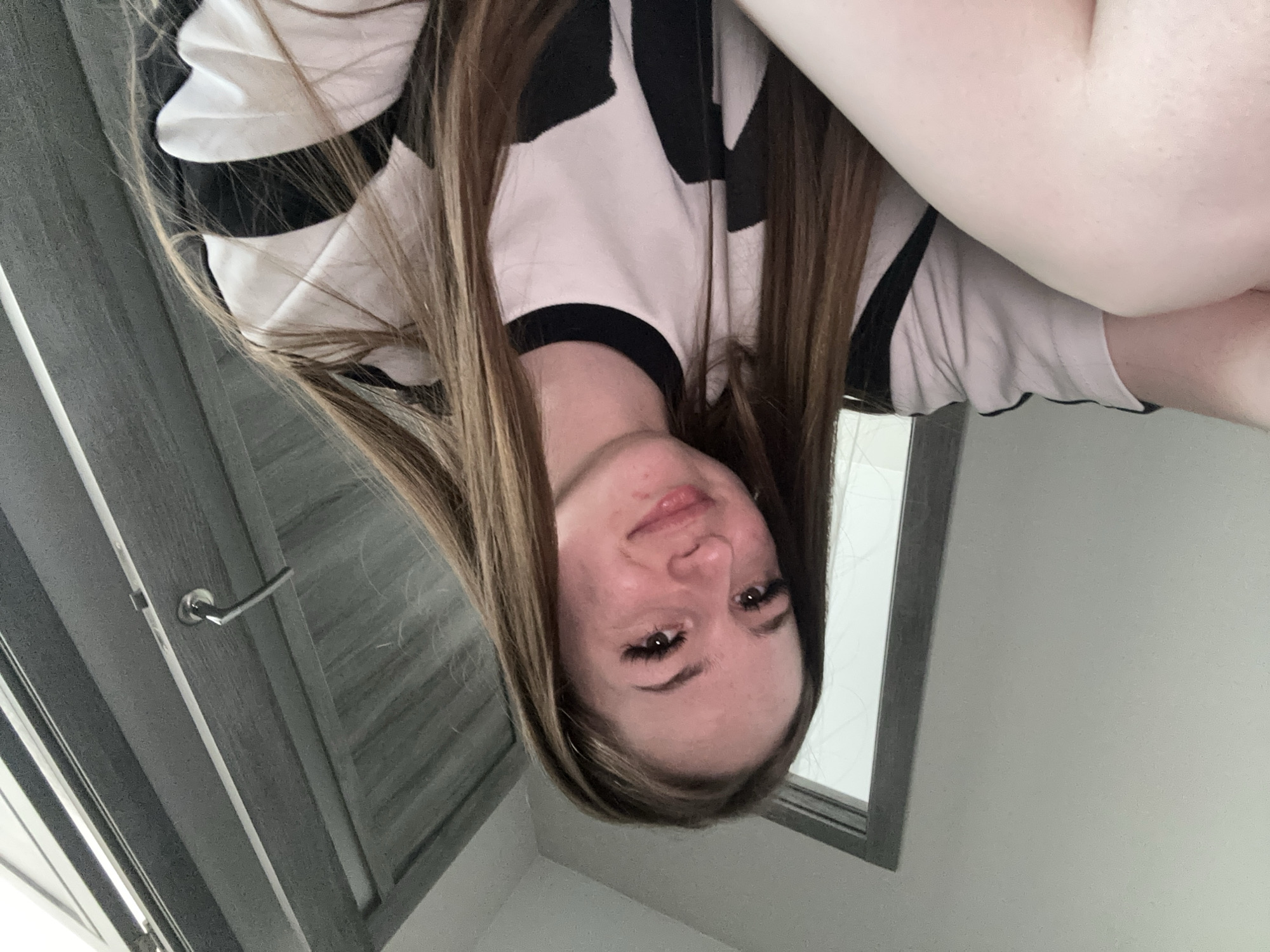 The image shows a young woman looking directly at the camera, with the image itself rotated 90 degrees clockwise, making her appear upside down. She has long, straight brown hair with lighter highlights, which frames her face and falls over her shoulders. She is wearing a black and white patterned top, possibly a t-shirt, with a black trim around the neckline.
The woman has fair skin, dark eyes, and well-defined eyebrows and eyelashes. Her lips are closed in a slight, subtle smile, and she appears calm and pleasant. Her upper body is visible, with her arms partially in the frame towards the top right of the image (due to the rotation).
The background on the left side features a textured gray surface, which appears to be a door or a panel, with a silver door handle visible. On the right side, there is a plain white wall and a dark-framed window or mirror reflecting a light surface. The lighting is bright and even, suggesting an indoor setting, but the time of day is not clearly discernible. There is no visible text in the image.