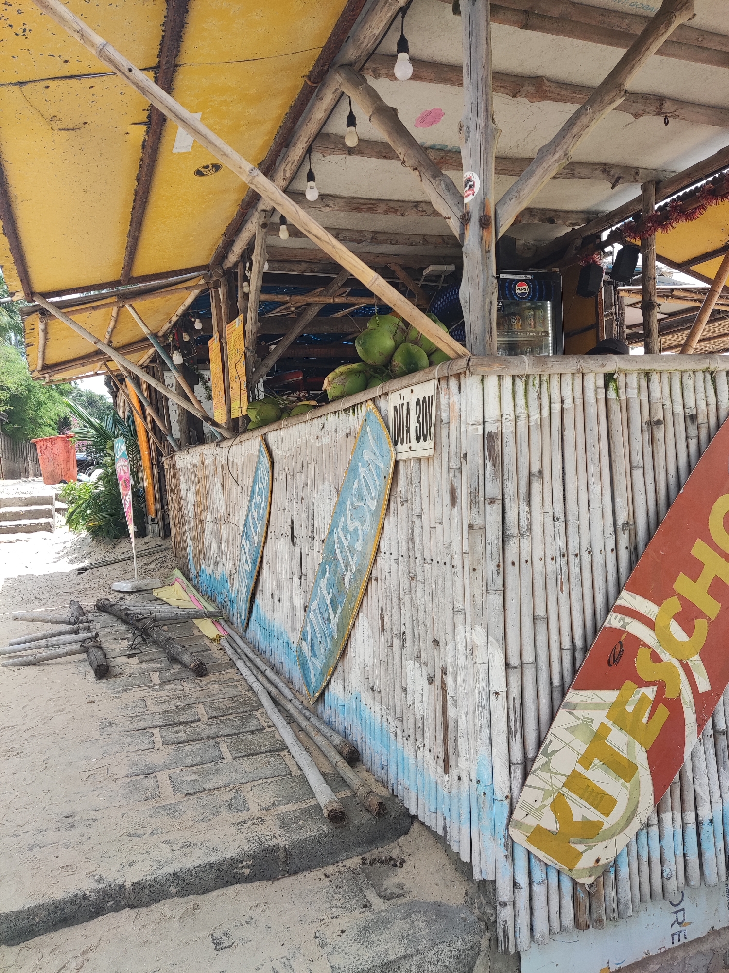 The image depicts an outdoor scene, likely a beachside establishment, constructed with natural materials. A weathered bamboo wall forms the facade of a bar or shop, with a yellow, tarp-like roof supported by wooden beams overhead. Several light bulbs are suspended from the beams. Coconuts are piled near the top of the bamboo wall. Leaning against the wall are two surfboards with text that appears to be in a foreign language. To the left, a pathway made of stone and sand leads away from the establishment, with more wooden poles lying on the ground. In the background, there is dense green foliage. The overall impression is of a rustic, possibly tropical, setting.