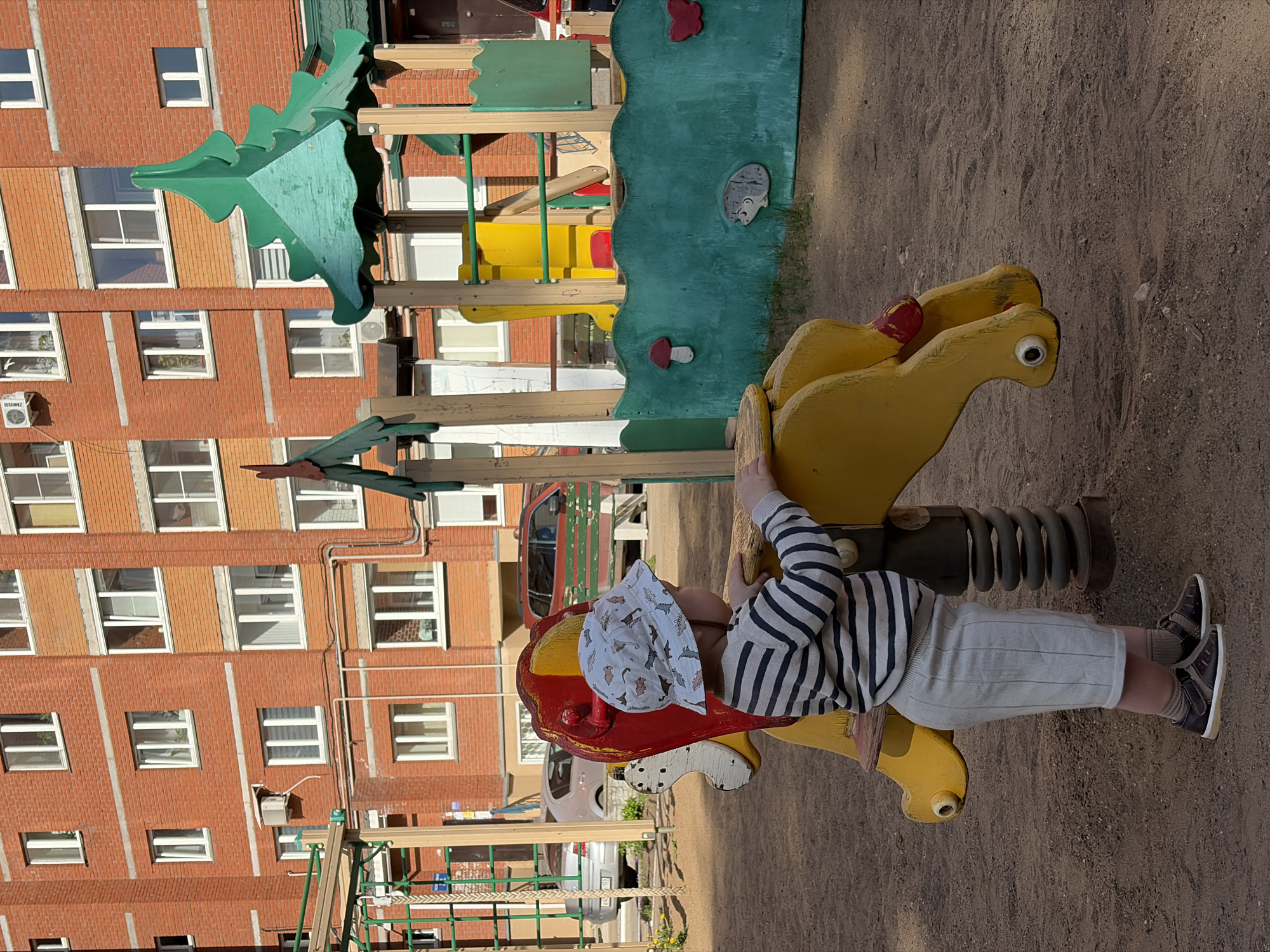 A young child is riding on a yellow and red spring-mounted playground horse in Chaikovsky, Russia. The child is wearing a striped shirt, light-colored shorts, and a patterned hat. Behind the horse, there is a colorful playground structure and a multi-story brick building. The ground is covered in dirt.
