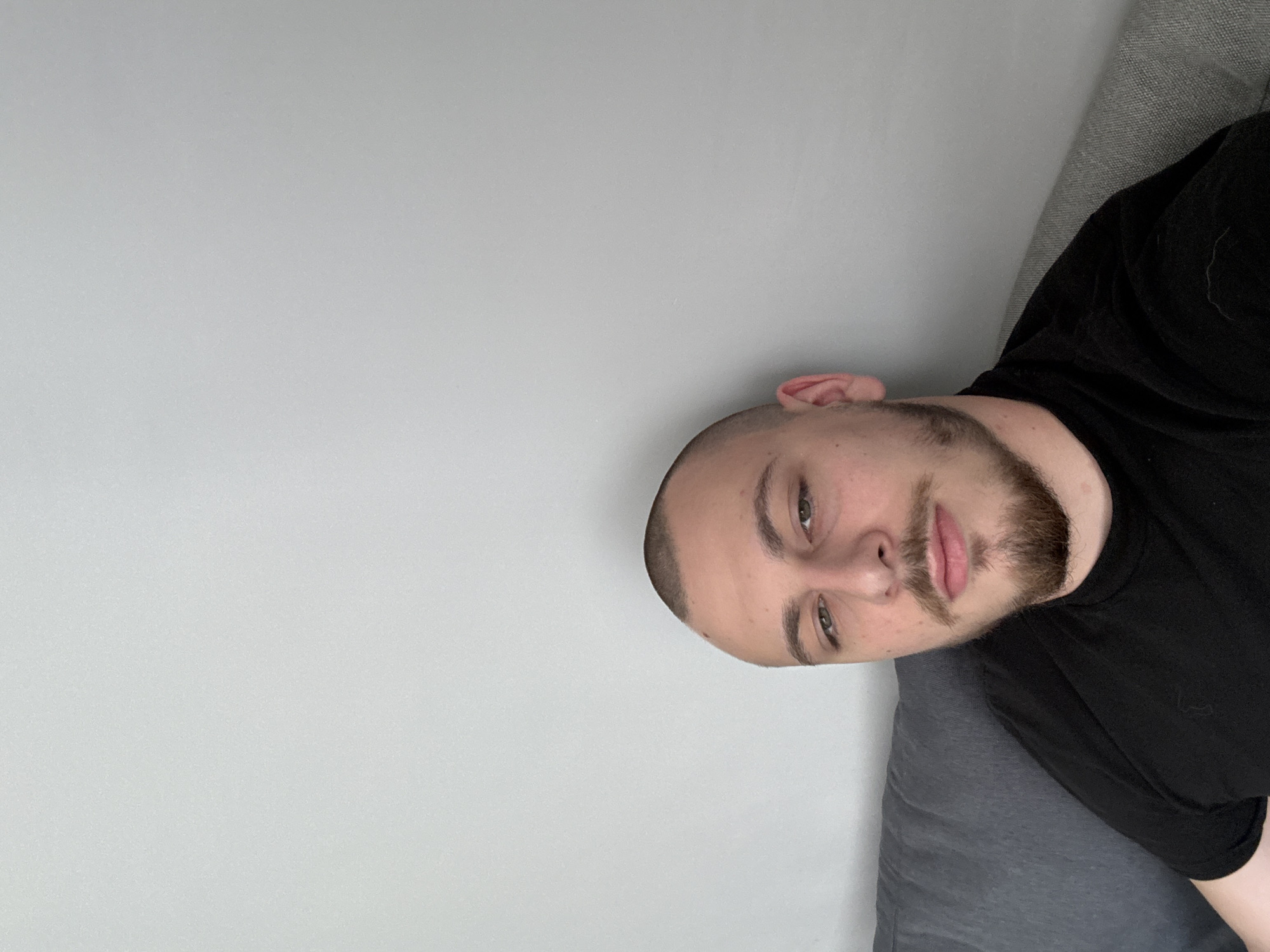 A medium, eye-level shot shows a man with a shaved head, light skin, and a beard. He wears a black t-shirt. He is lying on a gray fabric surface, likely a couch or cushion. The background consists of a light gray wall. The location is not discernible from the image.