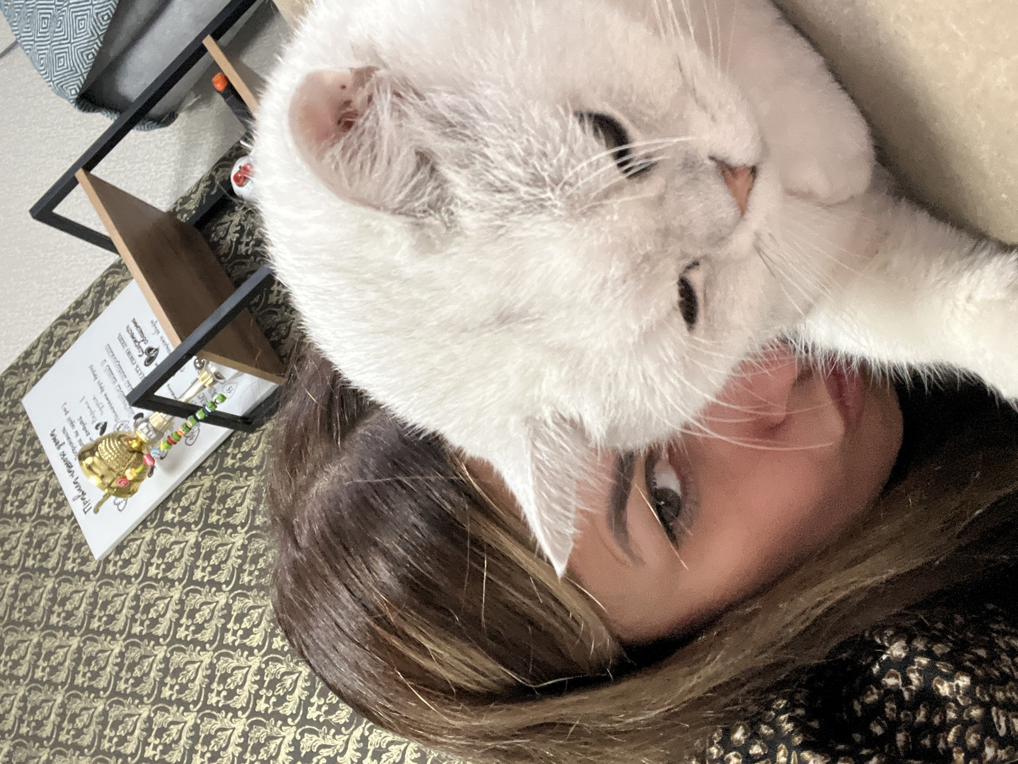An overhead, close-up, selfie-style shot shows the right eye and side of the face of a young woman with brown hair and dark brown eyes, who is looking down and to the left of the frame. To her right, a white cat with greyish undertones is partially obscuring the woman's face, with its head resting on her shoulder. The cat's eyes are closed, and its body is turned away from the camera. Behind the woman and cat, a black metal shelf unit is visible, with two wooden shelves. On the upper shelf, a white notepad with handwritten text and a small golden ornament with colorful beads is placed to the left. The background wall behind the shelf has a brown and gold patterned wallpaper. The scene appears to be indoors.