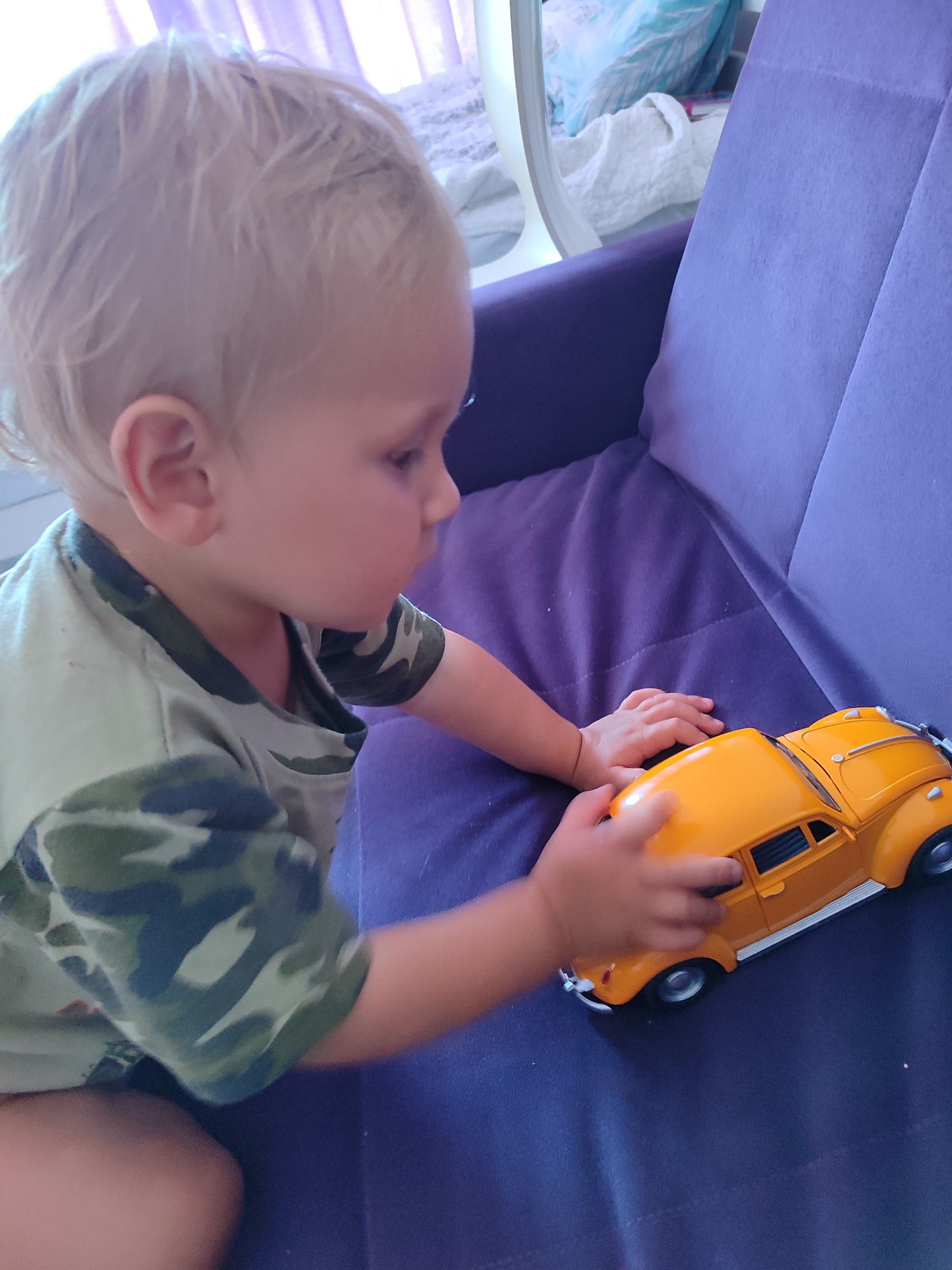 A blonde child sits on a purple sofa, playing with a yellow toy car. The child is wearing a camouflage t-shirt and is viewed from the side. The background shows a blurry image of curtains and possibly other furniture. The scene appears to be indoors. The image does not provide any specific information about the city of Sochi, Russia, or any other location details.
