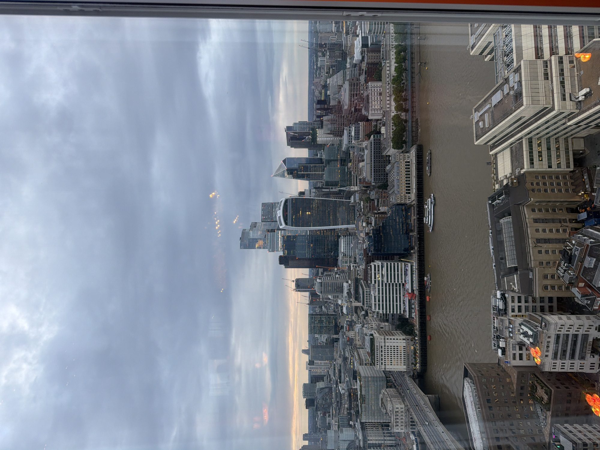 This is an aerial photograph of the London skyline taken through a window. The perspective is from a high vantage point, looking out over the River Thames towards the City of London financial district. The sky is overcast with grey clouds, and the light suggests it is either early morning or late evening, with the sun's last rays or first rays visible on the horizon as a faint orange glow.
The City of London, also known as the Square Mile, is visible in the middle ground, characterized by its dense cluster of modern skyscrapers and older buildings. Notable modern structures include the distinctive Gherkin building and other contemporary office blocks with glass facades and geometric designs. Cranes are visible among the buildings, indicating ongoing construction and development.
The River Thames flows through the foreground and middle ground, a wide, murky brown body of water. A bridge, likely London Bridge or a bridge nearby, spans the river on the left side of the image, with traffic visible on its surface. Several boats and barges are on the river, including a white passenger boat and a long, dark barge.
In the foreground, closer to the viewer, are more buildings, some appearing to be office blocks with flat roofs. Some of these foreground buildings have visible lights on, and decorative orange lighting fixtures are noticeable on the roof of one building. The overall impression is a view of a bustling, modern metropolis under a subdued, cloudy sky. The window frame, with a slight orange tint, is visible on the right edge of the photograph, indicating it was taken from inside a building.