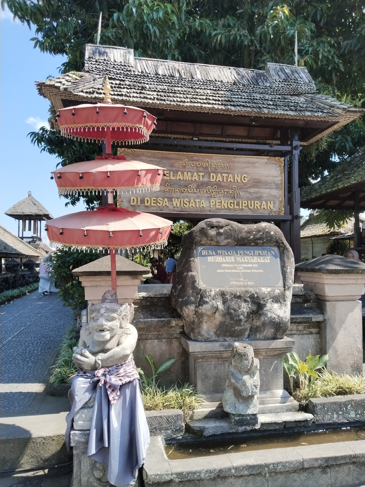 The image depicts the entrance to Penglipuran Village, a traditional village in Bangli, Bali, Indonesia. In the foreground, a stone statue of a Balinese guardian figure is adorned with a sarong and a sash. To the right of the guardian statue, a smaller stone figure kneels in a water feature. Behind the statues, a large stone serves as a plaque inscribed with text, indicating "DESA WISATA PENGLIPURAN BERBASIS MASYARAKAT" which translates to "Penglipuran Ecotourism Village Based on Community." Above this, a wooden sign with golden lettering reads "SELAMAT DATANG DI DESA WISATA PENGLIPURAN," meaning "Welcome to Penglipuran Ecotourism Village." The entrance is further decorated with three tiered red ceremonial umbrellas, characteristic of Balinese culture. The architecture features a traditional Balinese tiled roof supported by wooden posts. In the background, a pathway leads into the village, with more traditional structures and lush greenery visible. Several people are present, some walking along the path and others seated near the entrance. The scene is bathed in natural sunlight under a clear blue sky.