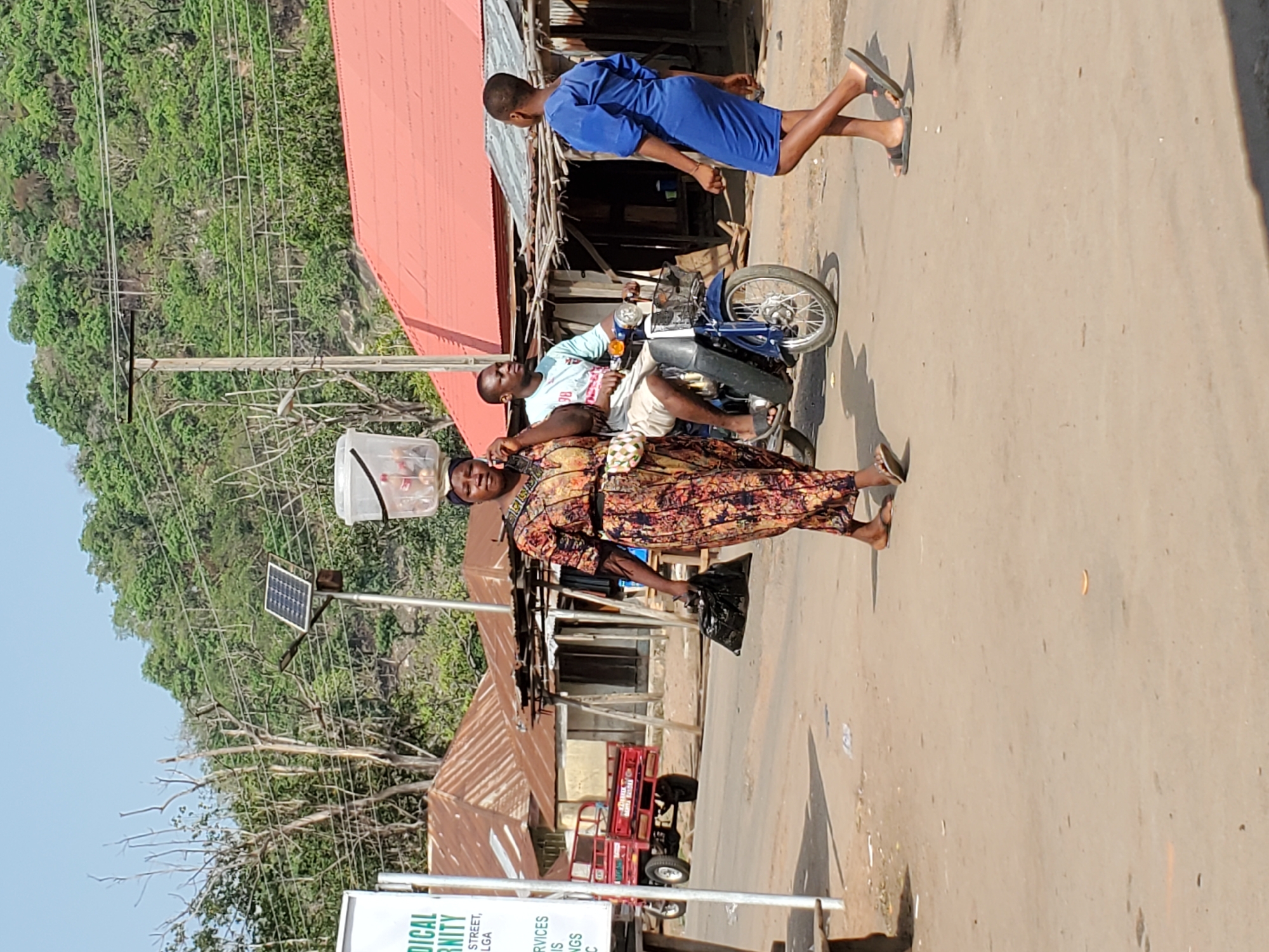 The image displays a lively street scene in Mararaba, Nigeria, captured during bright daylight. The main subjects are three individuals on a road. In the foreground, a woman in a long, vibrant patterned dress carries a white plastic container on her head and a black plastic bag in her left hand, suggesting she might be a street vendor or transporting goods. She wears sandals.
Behind her, a man sits on a blue motorcycle, looking towards the woman. Further into the background and to the right, another person in a blue tunic or dress walks away from the viewer, also wearing sandals.
The setting includes a dirt or unpaved road with distinct shadows, indicating a sunny day. The background features lush green hills or dense foliage. Several utility poles with power lines traverse the scene. Notably, one pole has a small solar panel attached, possibly powering a street light or other local infrastructure. Buildings with rusted, corrugated metal roofs are visible along the road, and a red three-wheeled cart or tricycle is parked nearby.
A white signpost in the lower left corner shows partial text, including "MEDICAL", "ERNITY", "STREET,", "LGA", "SERVICES", and "INGS". The scene captures everyday activity in a semi-urban or rural Nigerian environment.