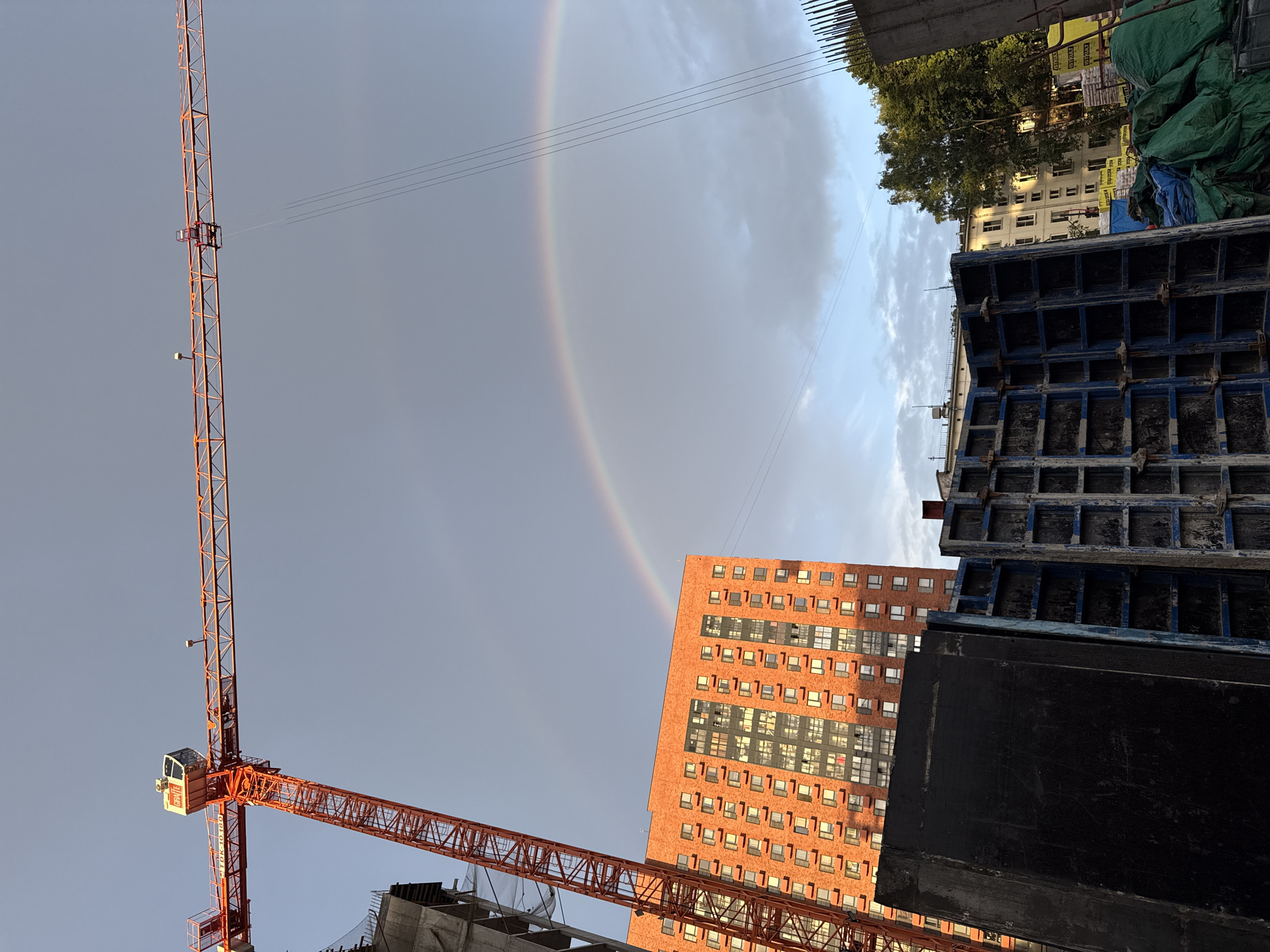 The image depicts a construction site in Moscow, Russia, with a prominent orange tower crane dominating the left side. A double rainbow arcs across a gray, cloudy sky, with the sun casting a warm glow on a modern, red-brick apartment building in the mid-ground. The building features numerous windows and balconies. In the foreground, construction materials are visible, including large black and blue formwork structures, and green tarpaulins covering objects. Other buildings and trees are visible in the background. The overall scene suggests an ongoing construction project under a sky transitioning from rain to clearing.