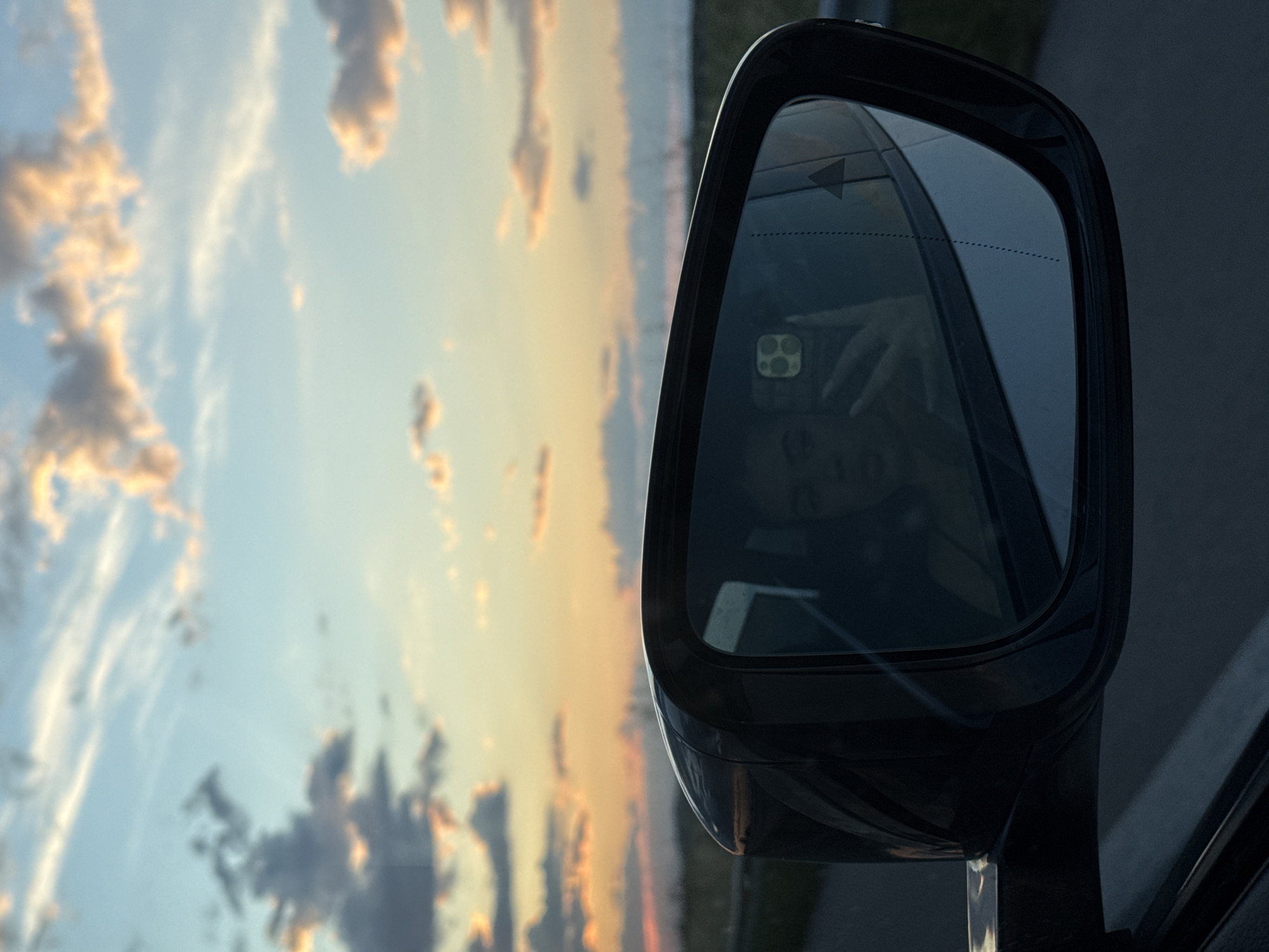 The image shows a car's side mirror against a backdrop of a sky with clouds during sunset, likely taken while driving. The mirror reflects a person holding a smartphone, presumably taking the picture. The location, Opalchenets, Bulgaria, cannot be determined from the visual content.