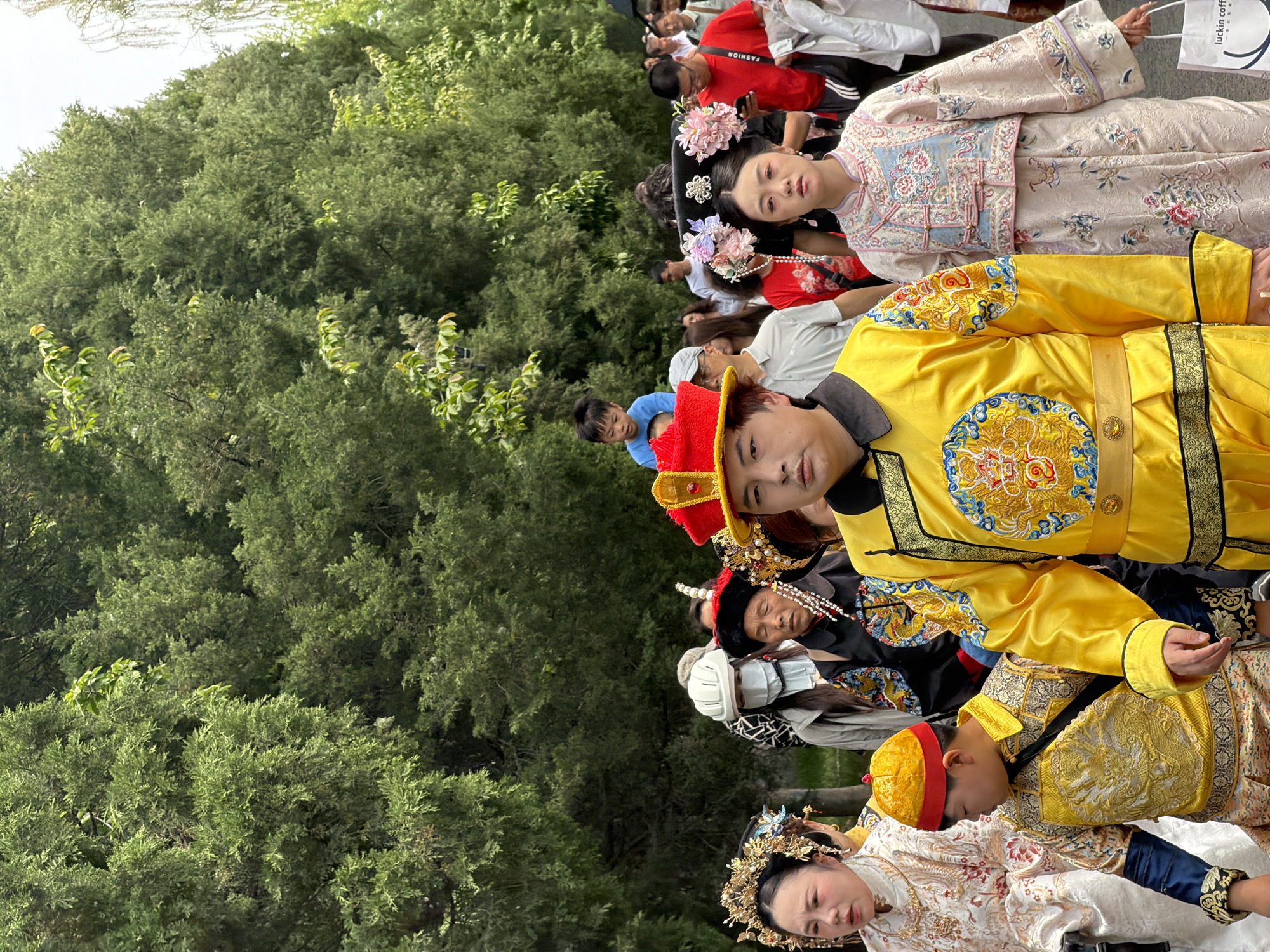 In Dongcheng District, China, a group of people, partially obscured by foliage, are dressed in traditional Chinese garments. Prominently featured are: a person in a bright yellow robe with gold embroidery and a red hat; a woman in a light pink and floral robe adorned with floral hair decorations; and other individuals in similar ornate attire. A few casually dressed onlookers are present in the background. A 'luckin coffee' branded bag is also visible. The environment includes green trees.