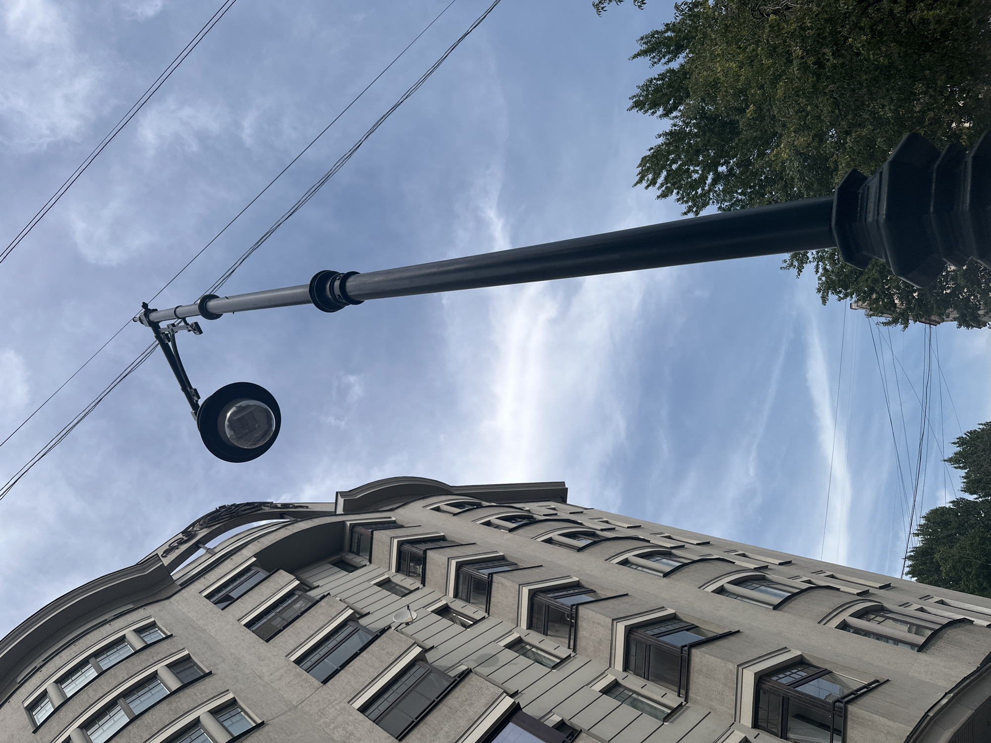 This is a low-angle, vertical outdoor photograph taken on a cloudy day in Moscow, Russia. The image features a gray, multi-story building with a curved facade on the left side, characterized by numerous windows with dark frames and small balconies. A tall, black street lamp with a circular light fixture stands to the right of the building, dominating the center of the frame. Overhead, several thin wires are stretched across the sky. In the background, the tops of green trees are visible, suggesting an urban park or residential area with greenery. The architecture and street lamp suggest a historic European city, consistent with Moscow.