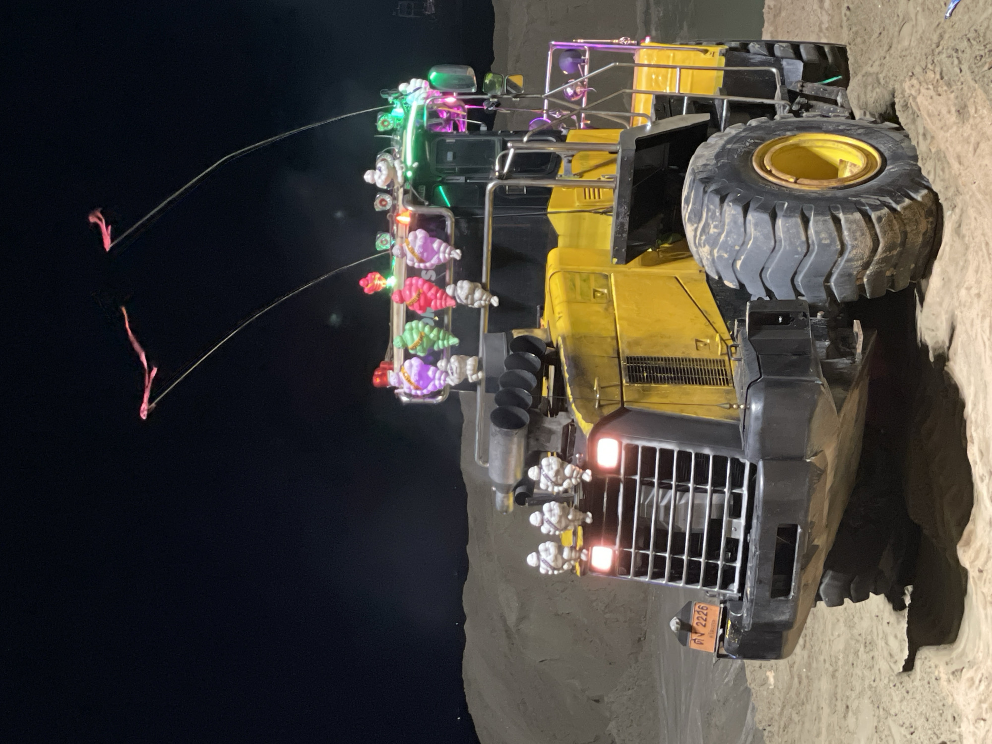 The image shows a large, yellow construction vehicle decorated with colorful lights and decorations. The vehicle appears to be parked on a dirt or sandy surface, possibly in a construction site or open area near a sand dune. A Thai license plate that reads "ภม 2226" is visible on the front of the vehicle. The vehicle is decorated with strings of lights and ornaments, including white marshmallows, which add a festive atmosphere to the scene. The image was taken at night, with the background appearing dark, suggesting it is likely that the image was taken in Pattaya City, Thailand.