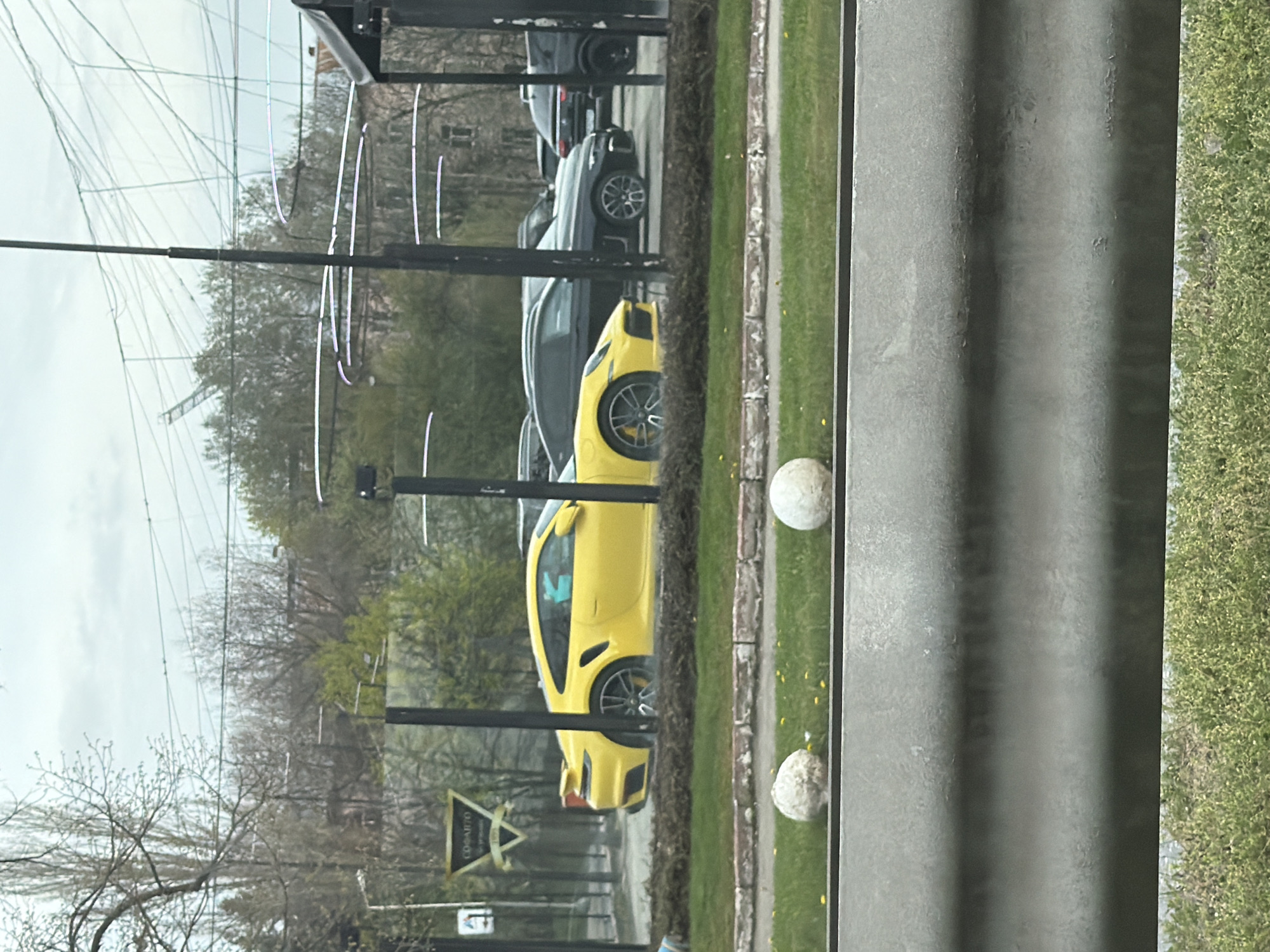 The image, rotated 90 degrees clockwise, captures an outdoor urban or suburban scene under an overcast sky.
In the foreground, a bright yellow sports car, possibly a Porsche, is prominently featured, facing right. It has multi-spoke wheels and visible brake calipers. Behind it, several other dark-colored cars, including SUVs and sedans, are partially visible.
To the right of the cars, a strip of green grass borders a paved area or sidewalk. On the grass, two white, spherical, textured objects, resembling decorative concrete spheres or large golf balls, are visible. Small yellow specks, possibly dandelions, dot the grass.
The left side of the image reveals numerous black poles, some supporting a large, fine-meshed net, suggesting a golf driving range or similar facility. Beyond the net and poles, a mix of trees, some bare and some with emerging green leaves, indicates a late winter or early spring setting. A building is faintly visible in the distant background. Overhead, various wires or power lines crisscross the sky.
A black and gold triangular sign is partially visible in the mid-ground, displaying the Cyrillic text "СОФАВТО" (SOFAVTO), which is consistent with the provided location context of Sofia, Bulgaria. The lighting is soft and diffused, characteristic of an overcast daytime.