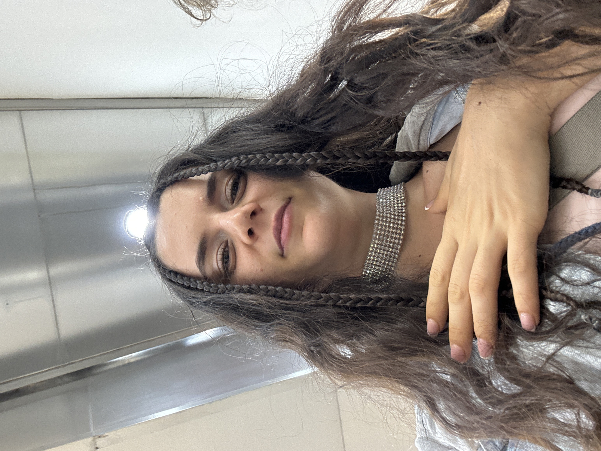 A young woman with long, dark brown hair braided on either side of her face is looking towards the camera. She is wearing a silver choker necklace and a gray top. Her left hand is resting on her chest. The background appears to be a plain, possibly white or light gray, wall and surface. The location is within Phu Thuy Ward, Vietnam.