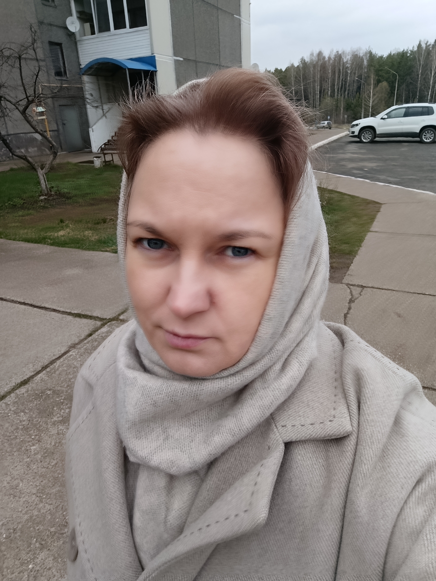 A woman with light skin and blue eyes, wearing a light grey headscarf and coat, poses for a selfie. The background includes a grey apartment building, a barren tree, a parking lot with a white SUV, and a line of trees in the distance. The location is Марковский, Russia.