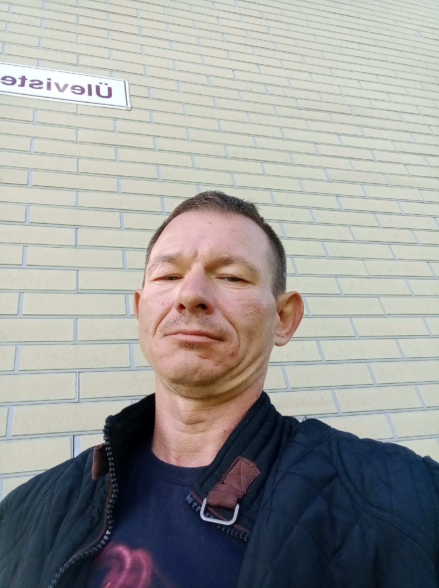 A man stands in front of a pale yellow brick wall. A white sign with purple lettering is visible on the wall in the upper left corner. The man is wearing a dark quilted jacket over a dark t-shirt. The t-shirt has a graphic design on it. The sign on the wall reads "Tapa Ühisgümnaasium", which translates to "Tapa United High School" and indicates the location in Tapa, Estonia.