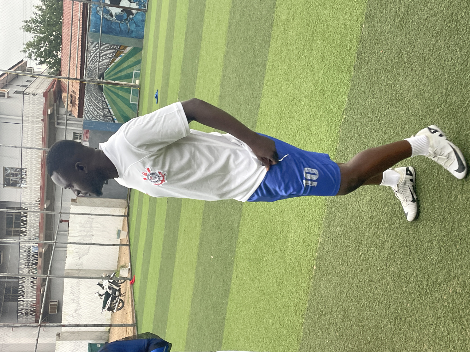 The image depicts a young man standing on an artificial turf field. He is wearing a white t-shirt with a crest and blue shorts with the number "10" on the left leg. He is also wearing white socks and white athletic shoes with black accents. The field appears to be a soccer field, with green turf marked with darker green stripes. In the background, there is a fence, a building with balconies, and a mural painted on a wall depicting a soccer stadium tunnel. There are also a couple of motorcycles parked behind the fence. The lighting suggests it is daytime. The provided context indicates this scene is located in Jos, Nigeria.