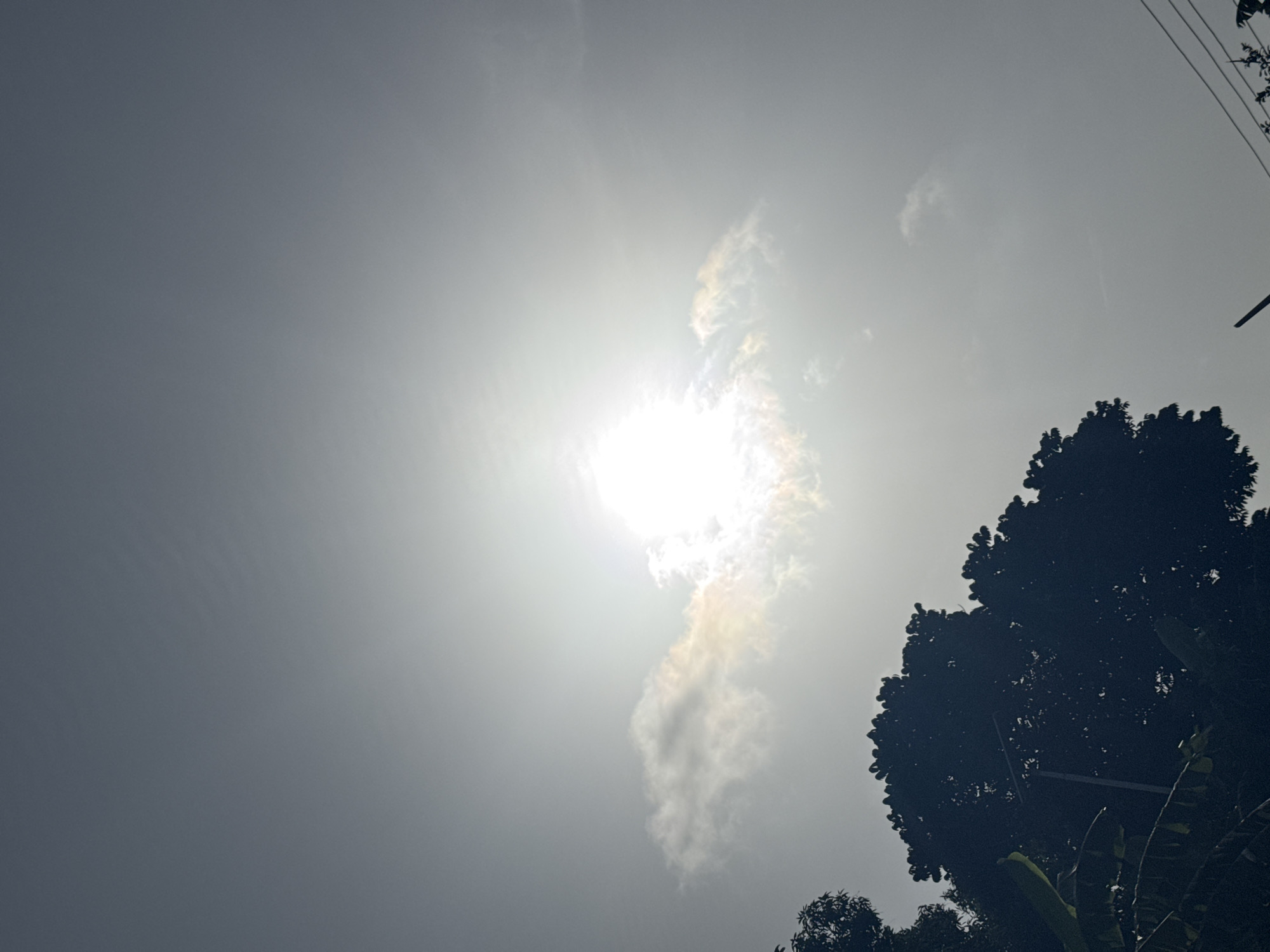 The image captures a daytime scene in Eziudo, Nigeria, under an overcast sky. The bright sun is partially obscured by clouds, casting a hazy light across the sky. In the lower right corner of the frame, the dark silhouette of a tree is visible. Power lines can also be seen in the top right of the image.