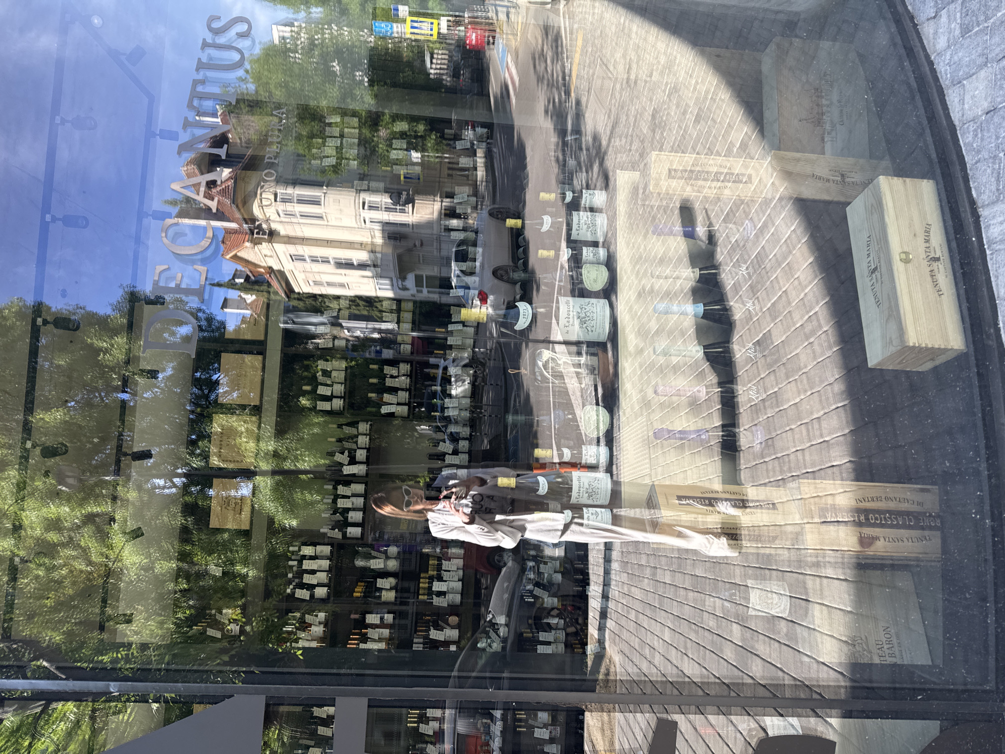 The image shows a storefront with the word "DECANTUS" prominently displayed. There is a reflection of a building and trees in the glass. Inside, shelves are stocked with numerous bottles of wine. A person, likely taking a photo, is reflected in the glass. Wine boxes with labels such as "TENUTA SANTA MARIA" and "AMARONE CLASSICO RISERVA" are visible. The wine "de Ladoucette" can also be seen. The city is Sofia, Bulgaria.