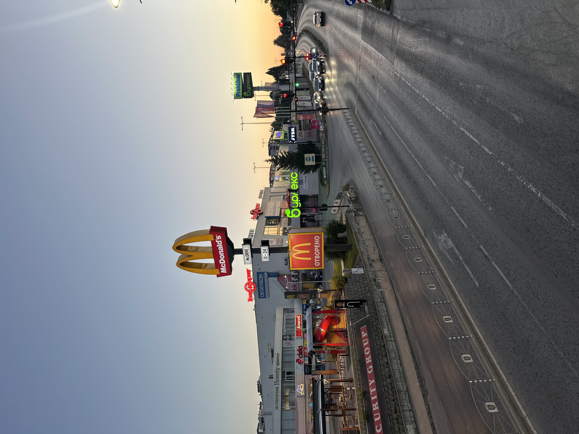 The image depicts a street scene in Veliko Tarnovo, Bulgaria, at dusk. A large McDonald's sign with its iconic golden arches is prominently displayed. Adjacent to it, a McDonald's restaurant with outdoor seating and a playground is visible. Several other businesses are situated in the buildings lining the street, including Decathlon, dm, Ciela, Komscel, and Kik. A large green sign with Bulgarian text, possibly advertising "Praktiker" and "Sydlex," stands out. The street is a multi-lane road with cars driving, and traffic lights are visible, indicating an intersection. The sky transitions from a clear blue to a warm, golden hue near the horizon, characteristic of sunset. A decorative metal fence with shield-like emblems lines the side of the road. The overall impression is of a commercial area on the outskirts of the city.