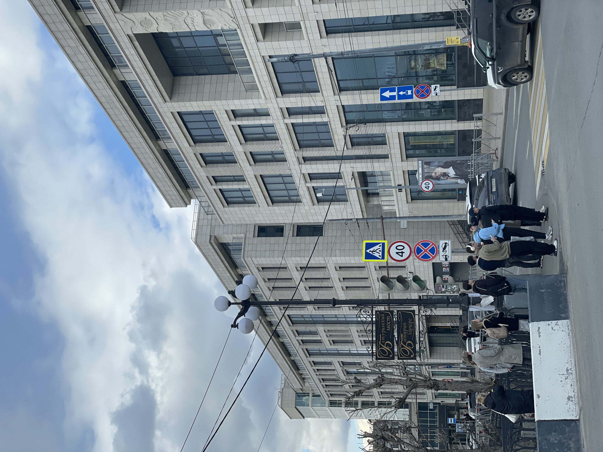 This image depicts a daytime street scene in an urban area of Kazan, Russia, under a partly cloudy sky. A large, modern-style building with a light-colored, possibly stone facade and many rectangular, reflective windows fills the background. Decorative elements are visible on the building's upper section.
On the sidewalk in the midground, a group of about five casually dressed people are gathered near a zebra crossing, appearing to wait to cross the street. Other pedestrians are visible further down the pavement. A dark car is partially visible parked near the curb, with another car behind it displaying a Russian license plate (partially visible "Н 152 КЛ 76").
Prominent street furniture includes a black lamppost with multiple globe lights, vertical traffic lights, and several road signs. These signs include a yellow diamond indicating a pedestrian crossing, a red circular speed limit sign displaying '40' (km/h), a blue rectangular sign with a white left-pointing arrow and car symbol, and a red circular 'no stopping' sign. A distinctive ornate black sign with gold Cyrillic text reads "Амурский Мост" (Amursky Most) and "Батуринские Коттеджи" (Baturin Cottages). An outdoor advertising board featuring a person's image is also present. Electrical wires crisscross parts of the scene. The overall impression is a typical, moderately busy city street.