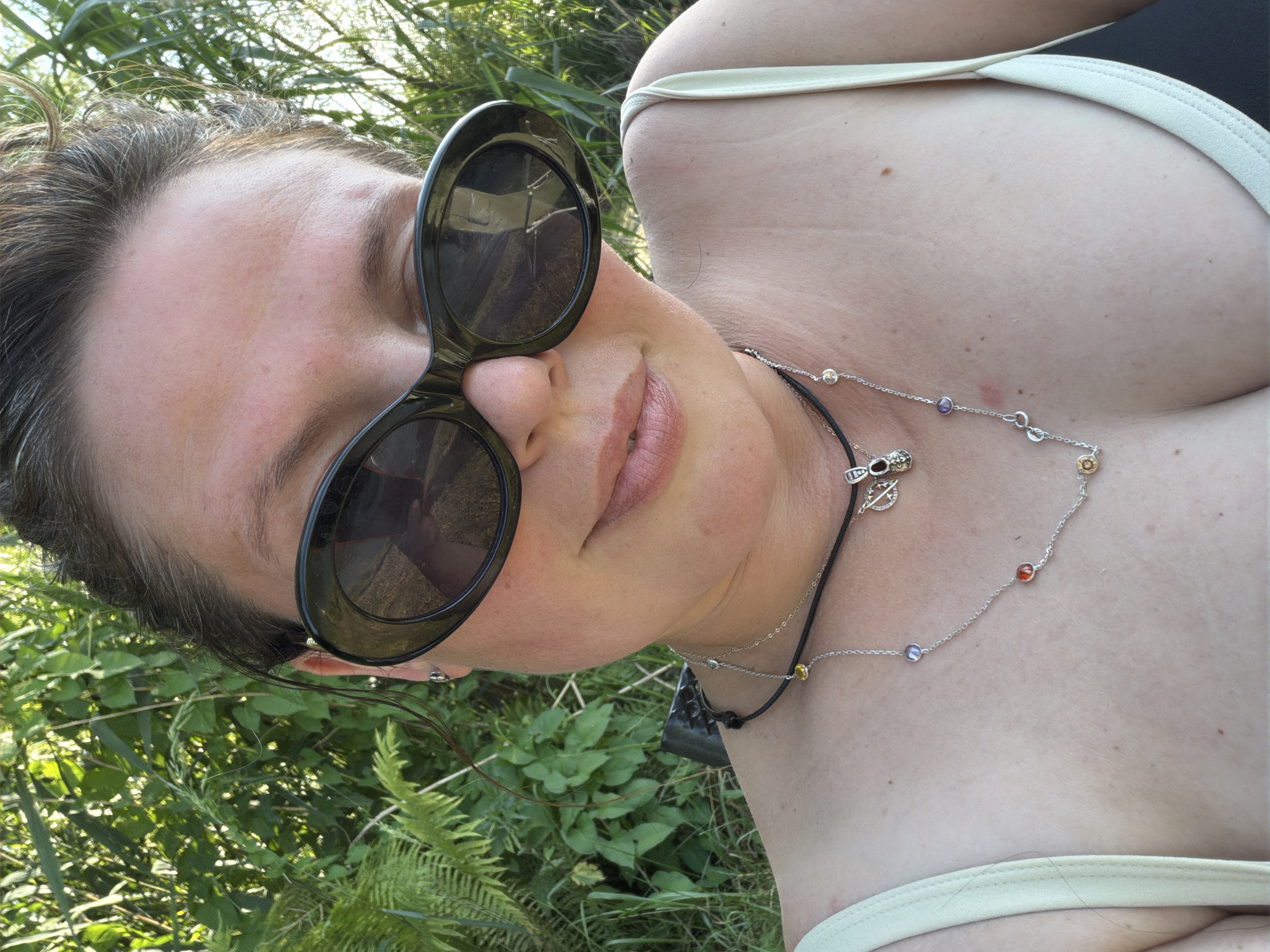 The media file shows a close-up, tilted shot of a person's face and upper body, outdoors. The subject is wearing large, dark-rimmed sunglasses and a light-colored strappy top. A silver necklace with colorful, small circular charms and a dark pendant is visible around their neck. The background consists of lush green foliage, suggesting a natural, possibly grassy or wooded, setting. Sunlight filters through the leaves, indicating it is daytime, likely with clear or partly cloudy weather. The person's head is tilted, and they appear to be looking upwards or to the side, with a relaxed or content expression suggested by their slightly parted lips. The overall impression is casual and relaxed, possibly taken during a summer day outdoors in Kamianka-Buzka, Ukraine. There is no visible text in the image.