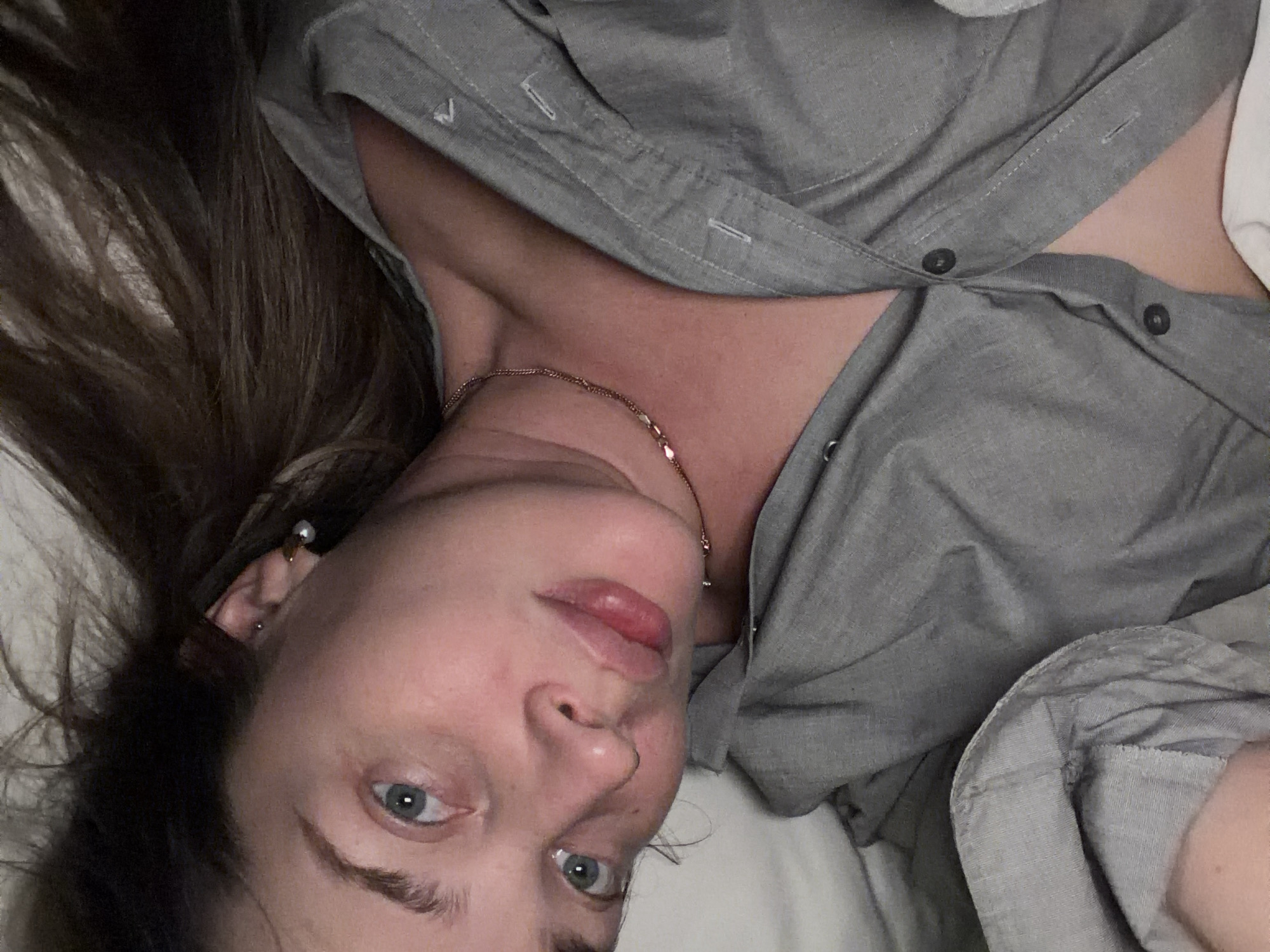 A woman lies supine against a white surface, possibly a bed. She is wearing a gray button-down shirt, partially unbuttoned, and a gold necklace. She has light skin, blue eyes, and brown hair. She has multiple ear piercings in her left ear. The image does not provide any details that would allow for the identification of a specific location in Moscow, Russia.