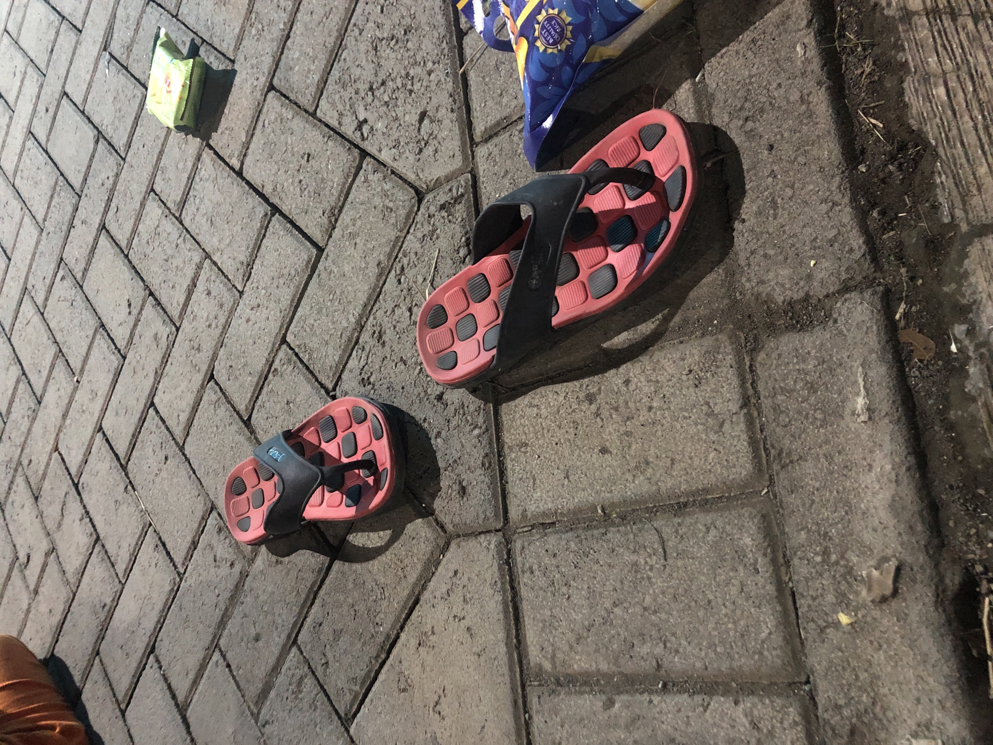The image shows a pair of pink and black flip-flops lying on a paved ground surface. The paving stones are rectangular and arranged in a herringbone pattern. In the background, a green packet is visible on the paving stones, and a portion of a person's orange clothing is in the upper left corner. To the right of the flip-flops, a blue and yellow object, possibly packaging, is partially visible. The scene appears to be outdoors, on a street or pathway, likely in Tenggilis Rejo, Indonesia, given the context.