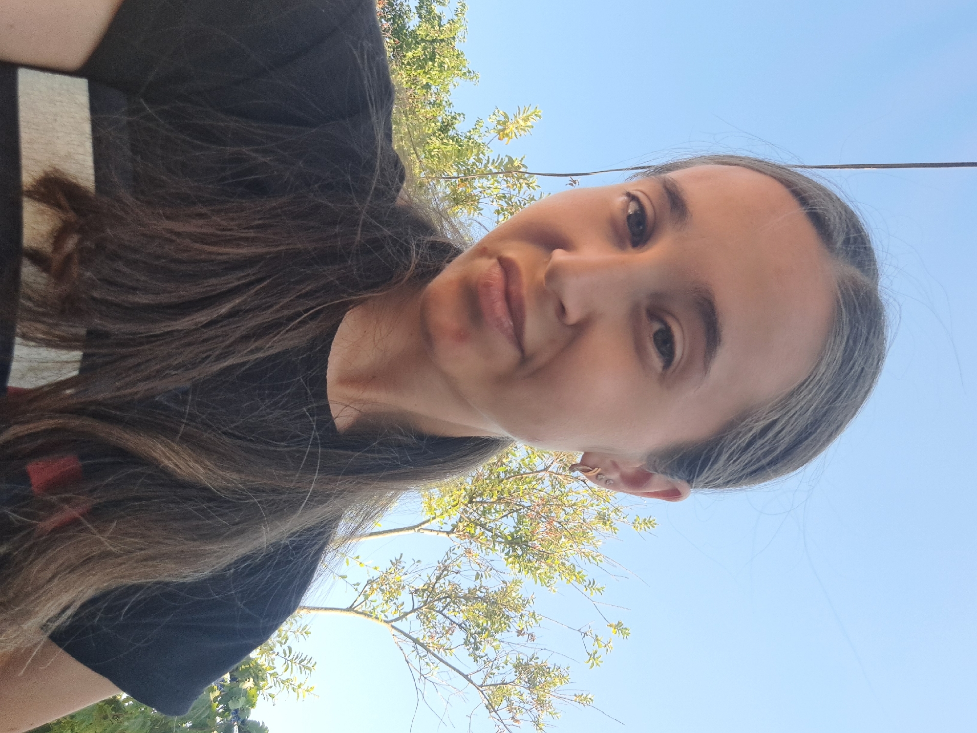 A young woman with long, brown hair pulled into a ponytail is photographed from the chest up. She is wearing a black t-shirt with horizontal stripes. Her face is turned slightly towards the camera, and she has a neutral expression. Behind her, a clear blue sky is visible, with green trees and branches. A thin wire hangs vertically in the background to the left of her head. The image appears to be taken outdoors, in a setting with natural light. There is no indication of the specific location within Vypasne, Ukraine.