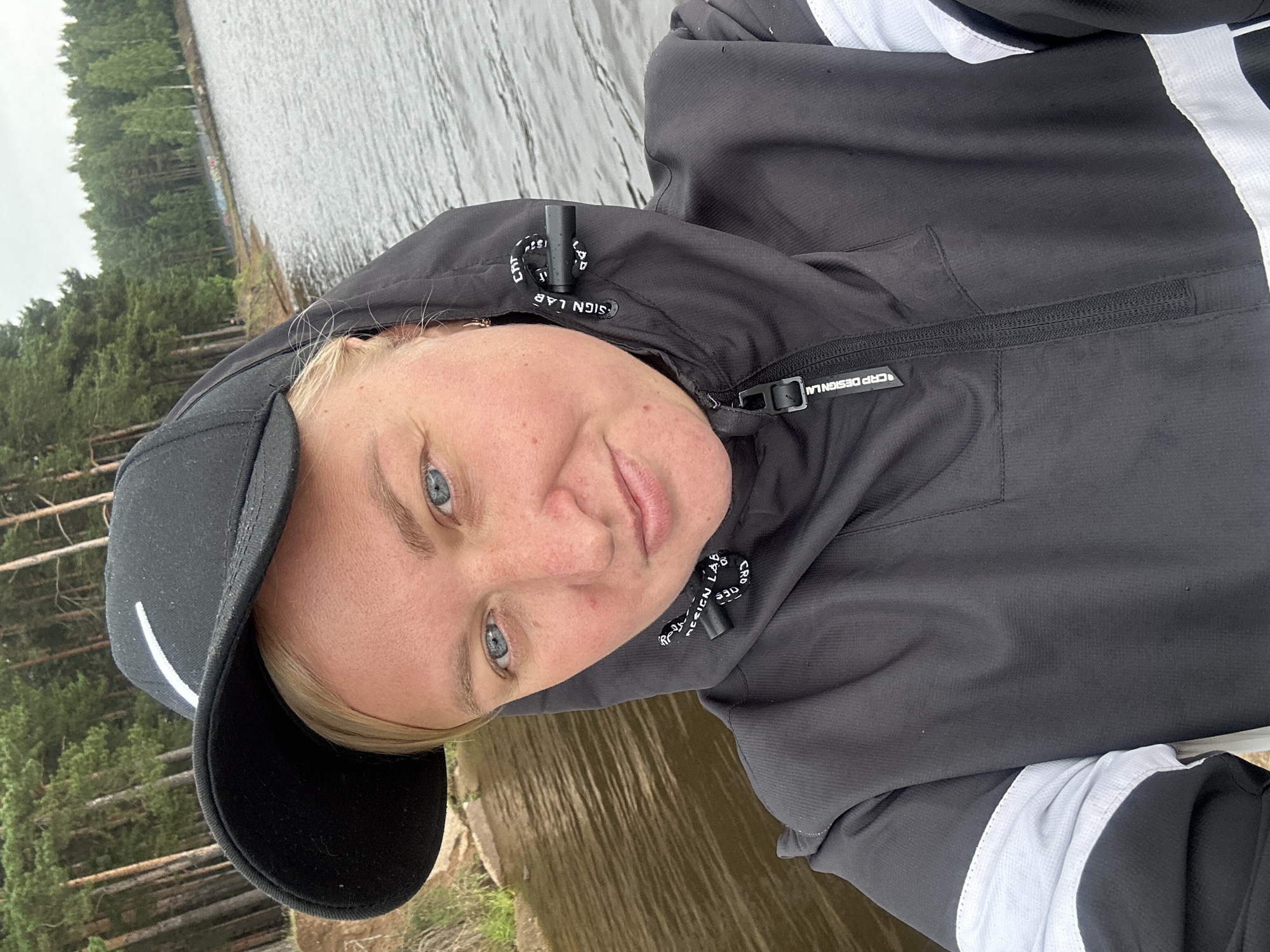 A portrait of a woman wearing a black baseball cap and a black hooded jacket with white accents on the sleeves. She has blonde hair and blue eyes and is looking directly at the camera with a slight smile. The woman is outdoors, with a body of water and a dense forest in the background. The water appears calm, reflecting the overcast sky. The trees are tall and green, suggesting a natural, possibly wild, setting. The overall atmosphere is relaxed and casual, possibly indicating a leisurely activity like hiking or enjoying nature. The image appears to have been taken during the daytime, under cloudy weather conditions. The provided location context of Chaikovsky, Russia, places the scene in a northern European environment, consistent with the depicted landscape.
Visible text includes "CRP DESIGN LAB" on the drawstring of the hood and on the zipper pull of a pocket.