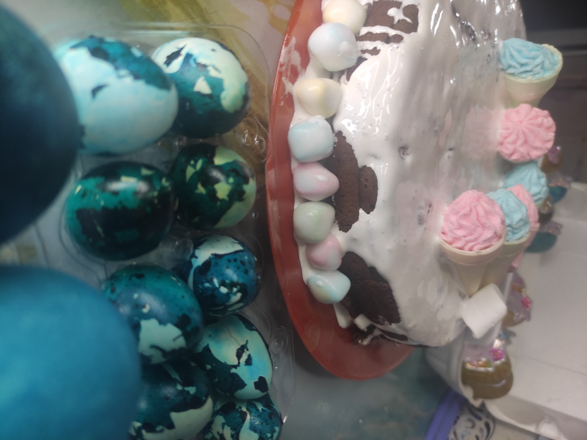 The image shows a top-down view of two food items: a container of dyed Easter eggs and a cake. The eggs are dyed in shades of blue and green, and some have a speckled or marbled appearance. They are arranged in a clear plastic container. The cake is decorated with white frosting and pastel-colored candy. It also features small, decorative ice cream cone figurines. The setting of Chaikovsky, Russia, is not evident in the image. There are no people, actions, interactions, or environmental details visible beyond the immediate food items.
