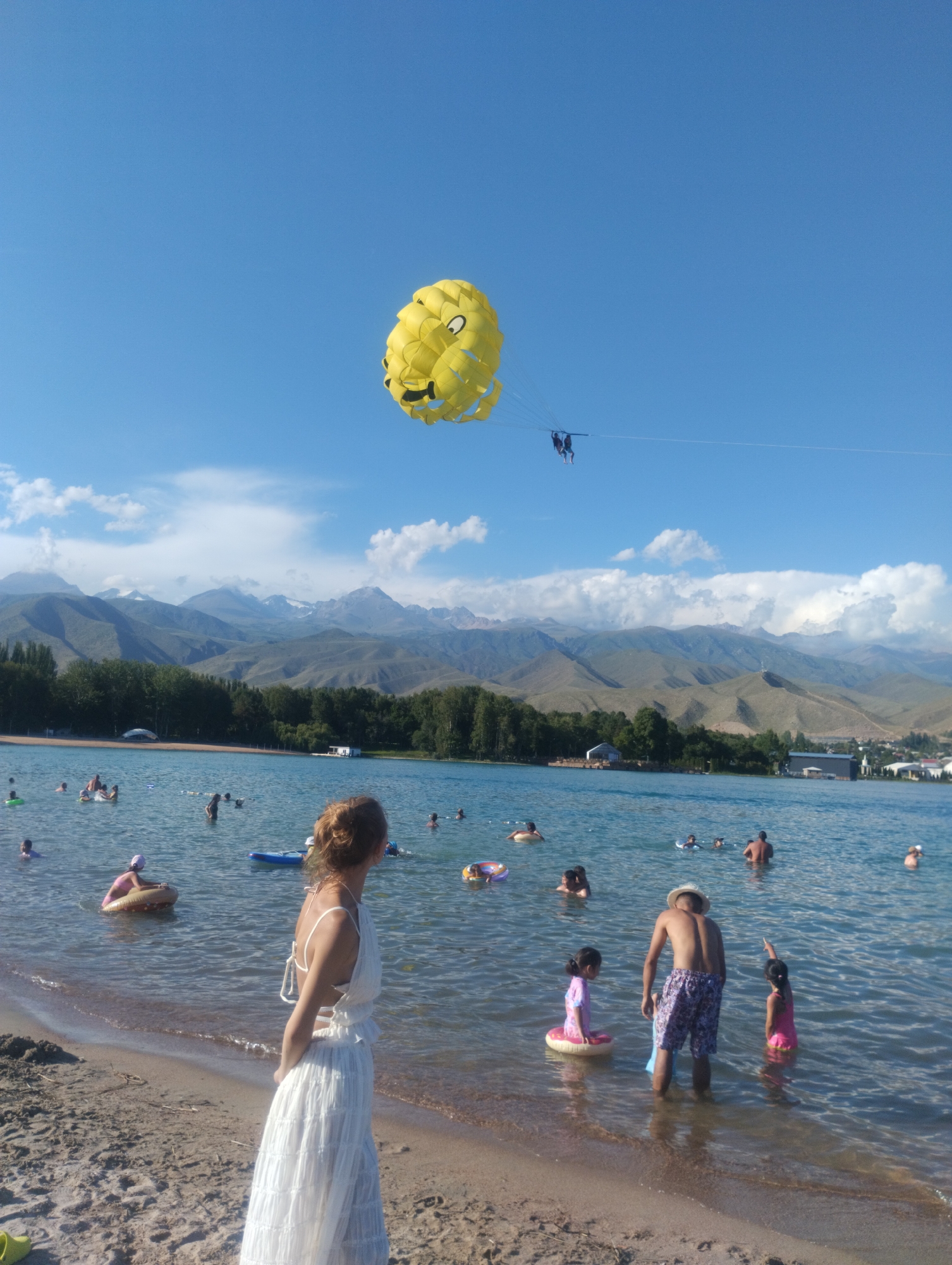 This image depicts a sunny day at Issyk-Kul Lake in Cholpon-Ata, Kyrgyzstan. A yellow smiley-faced parasail, with two people harnessed underneath, is visible in the clear blue sky. The lake is populated with numerous people swimming, wading, and relaxing on inflatable rings. In the foreground, a woman in a white dress stands on the sandy beach, looking out at the water. In the middle ground, a man wearing a straw hat and patterned shorts is standing in the water with two young girls. The background features lush green trees and a range of majestic, snow-capped mountains. Several buildings are scattered along the shoreline, and a small boat is docked near a building in the distance. The overall scene conveys a vibrant and enjoyable summer atmosphere.