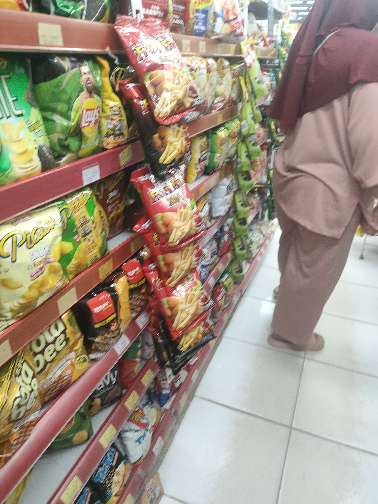 In Desa Banyumas, Indonesia, a woman wearing a maroon hijab and a light pink modest outfit stands in a grocery store aisle. She is facing away from the camera, with her feet clad in light pink sandals visible on the tiled floor. The aisle is stocked with shelves filled with various bags of chips and snacks. Prominent among the products are bags of Lay's potato chips, some featuring an image of a man, and other snack brands with colorful packaging. The shelves are red and the lighting suggests an indoor commercial environment.