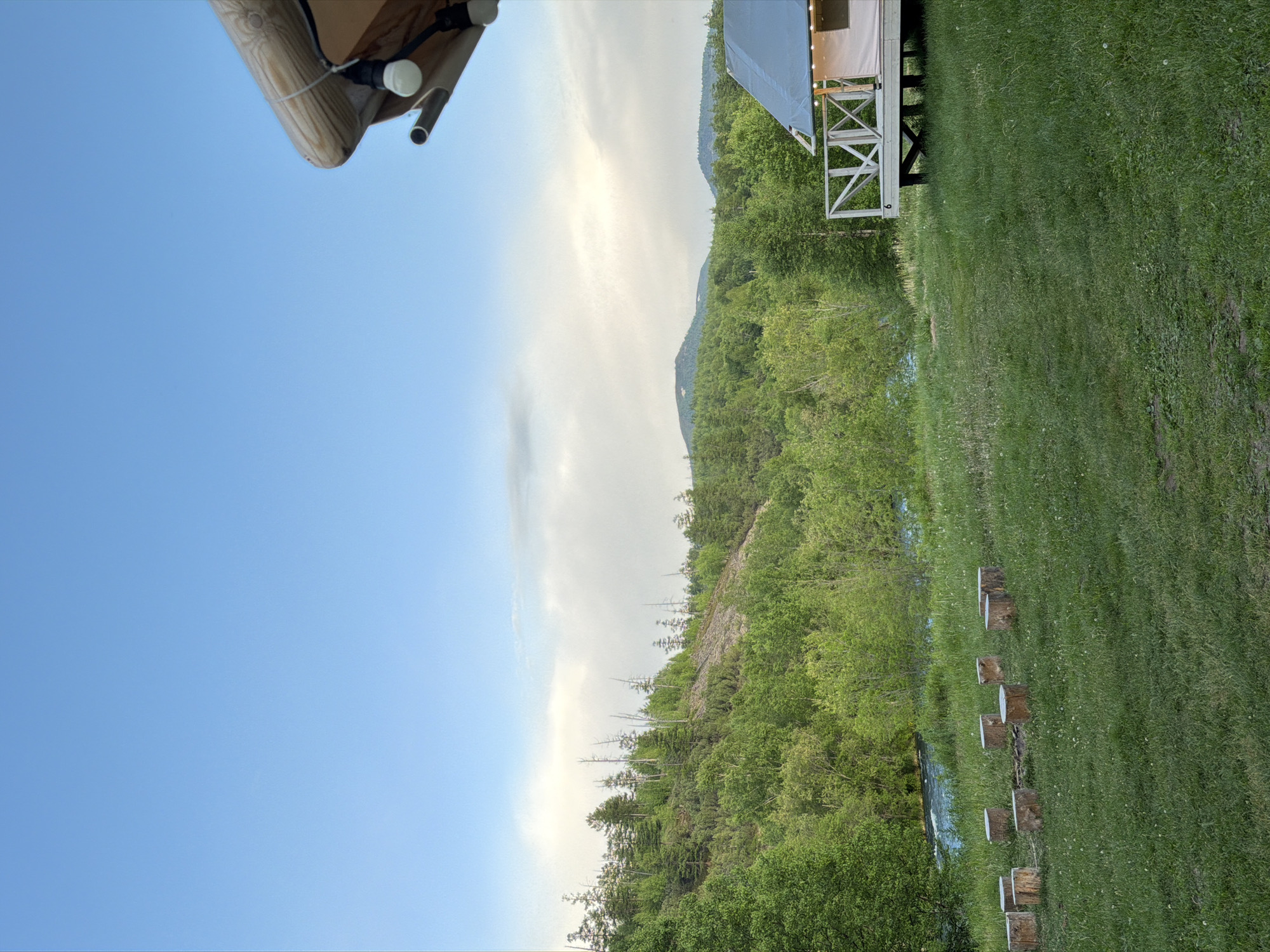 The image depicts a serene outdoor scene, likely a glamping site, set against a backdrop of lush green forest and rolling hills under a pale blue sky with scattered clouds. In the foreground, a grassy clearing features a circular arrangement of rustic wooden stumps, suggesting a fire pit area. To the right, a modern glamping tent, elevated on a wooden platform with a railing, is visible. String lights are draped over the tent's structure, and a number "6" is affixed to the railing. The middle ground is dominated by a winding river or body of water, flanked by dense, vibrant green trees. The background showcases a steep, tree-covered hillside with some sparser vegetation and dead trees, leading to distant, hazy mountains. The lighting suggests it is either early morning or late evening. The specific location in Esso, Russia, is suggested by the natural, wild landscape with a river and surrounding forests, characteristic of rural or wilderness areas in that region.