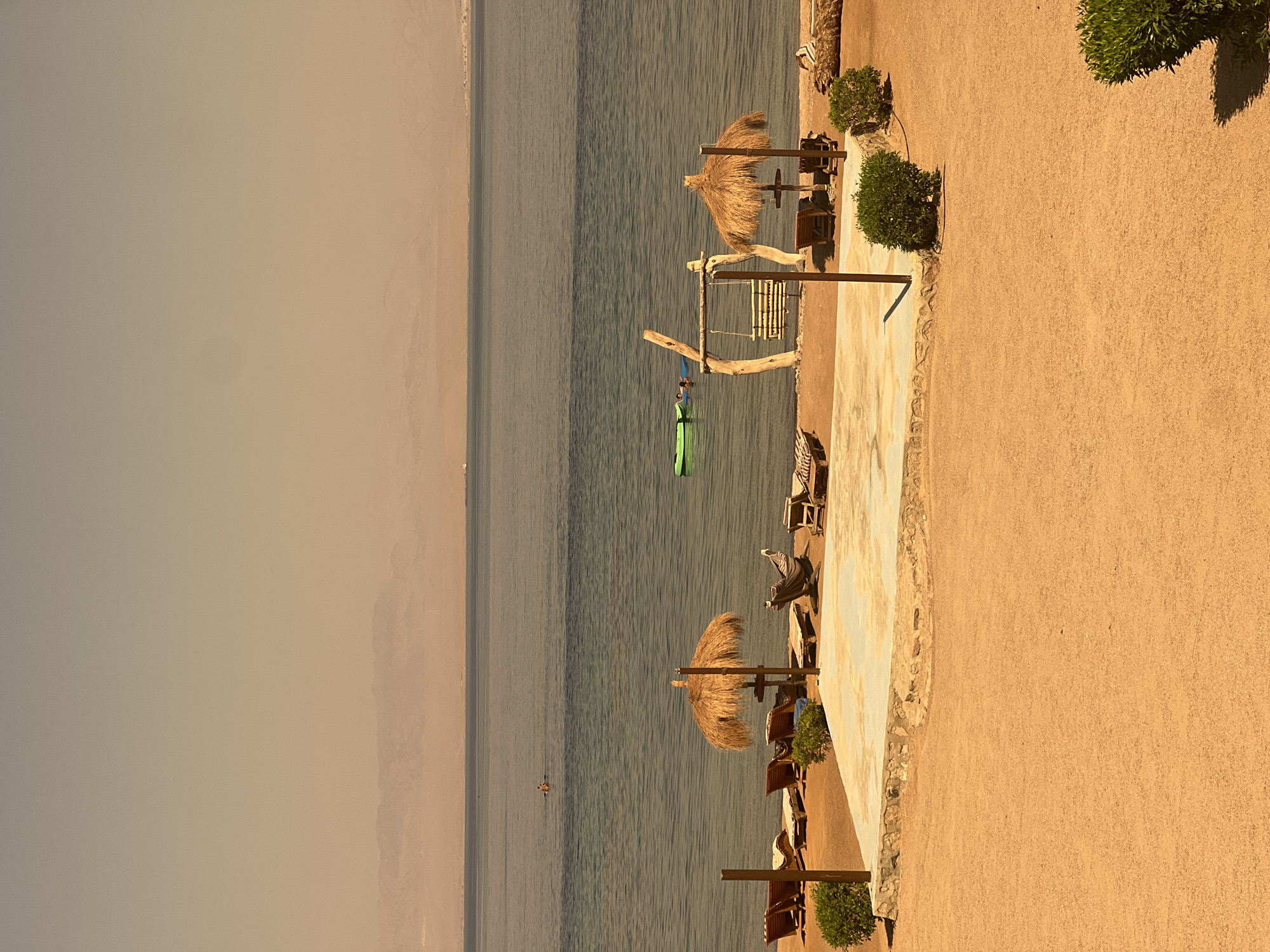This image depicts a tranquil beach scene in Nuweiba, Egypt. The foreground is dominated by a sandy beach with a raised concrete platform featuring a stone border. Several lounge chairs are scattered on the sand and platform, some positioned under thatched umbrellas. A wooden swing set is also present. The calm, blue sea stretches to the horizon, where a hazy, mountainous coastline is visible under a pale sky. Two individuals are in the water; one is on a green float, and another is further out on a paddleboard. The overall atmosphere is serene and uncrowded, suggesting a peaceful vacation setting.