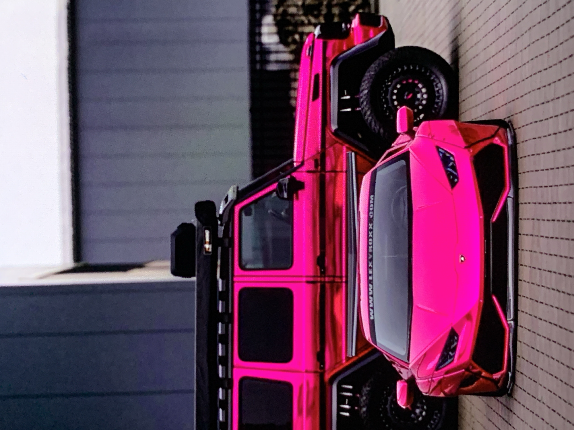Two vehicles are parked on a brick-paved surface in front of a building. The vehicle in the foreground is a bright pink sports car with a lowered stance and black accents. A banner across its windshield reads "WWW.LEXROXX.COM". Behind the sports car is a large, bright pink SUV with a matte black roof rack. The SUV has large, aggressive tires and black fender flares. The building behind the vehicles is modern, with dark gray vertical siding. The overall scene is outdoor and appears to be a car show or display.